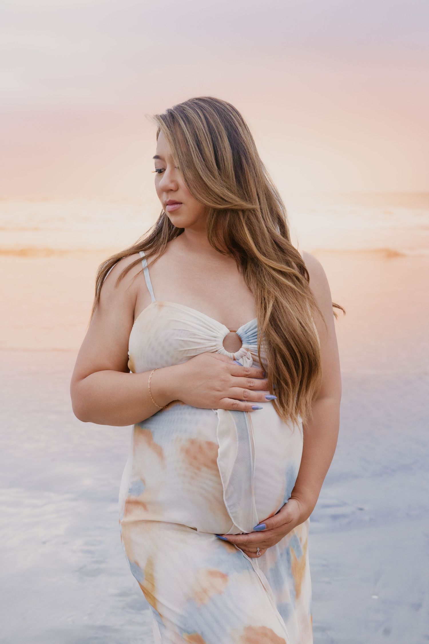 Pregnant woman in tie-dye dress cradling baby bump on beach at sunset with long flowing hair.