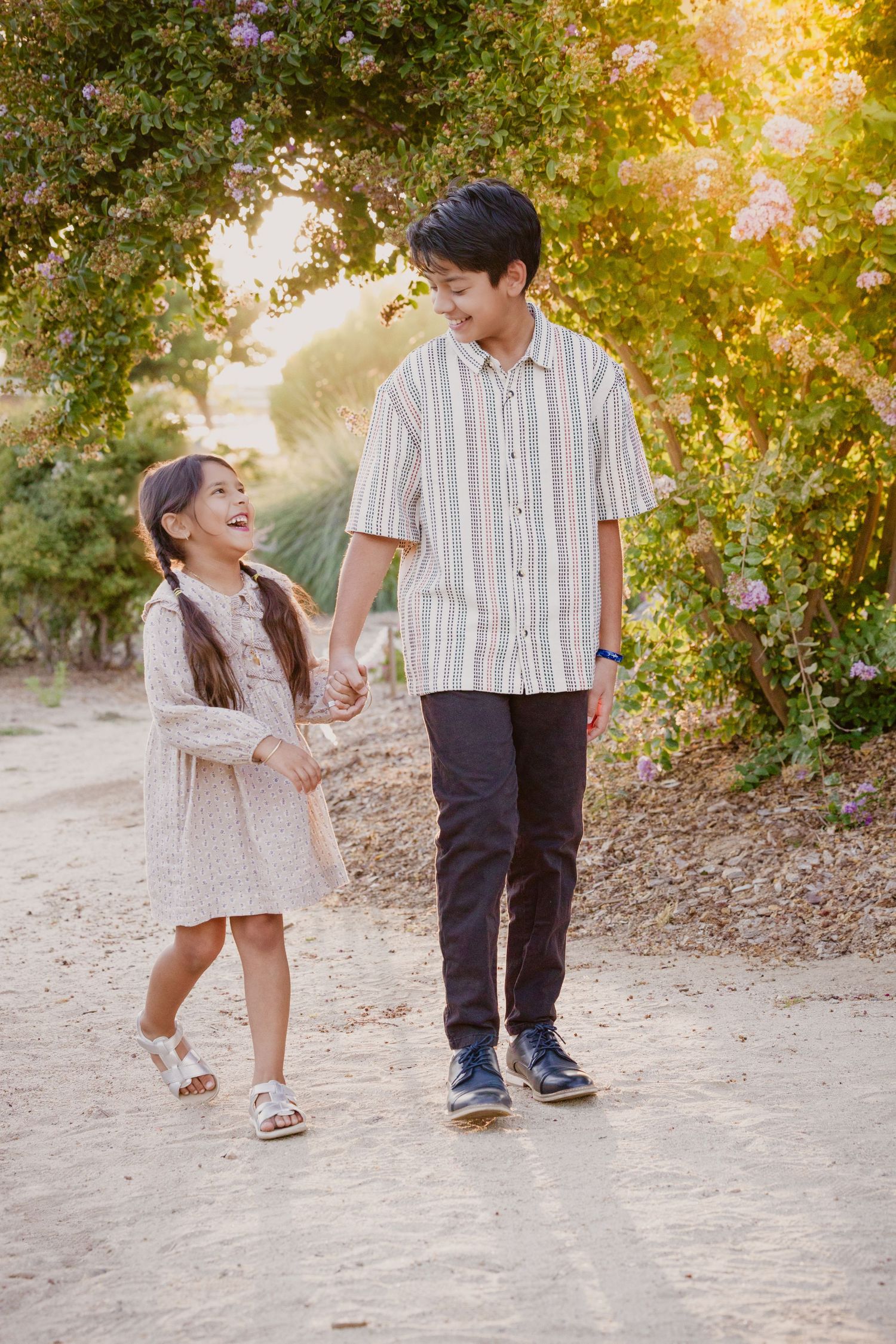 Two people walk hand in hand on a sunlit path surrounded by greenery during golden hour.