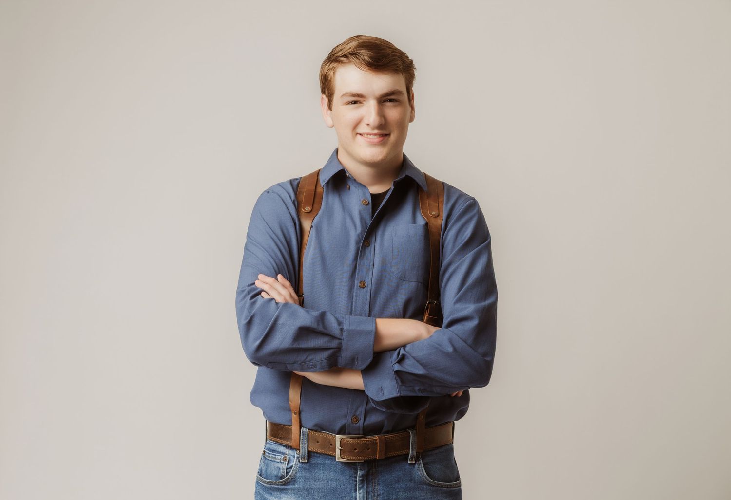 Smiling young man with arms crossed wearing blue shirt and suspenders against gray background.