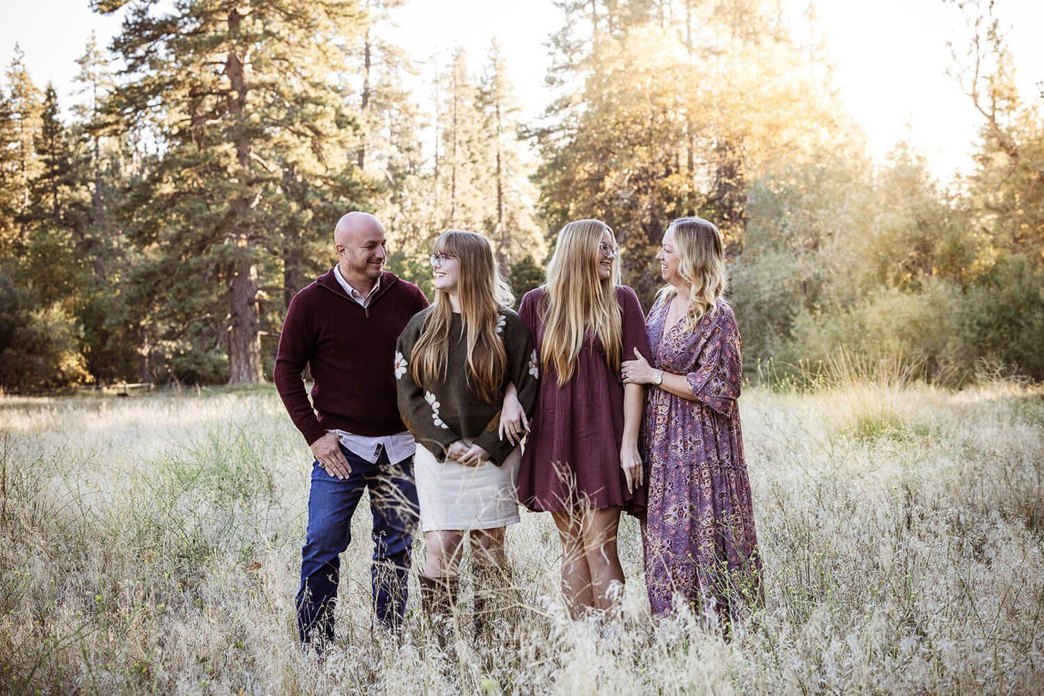 A family of four poses together in a sunlit meadow surrounded by tall grass and pine trees.