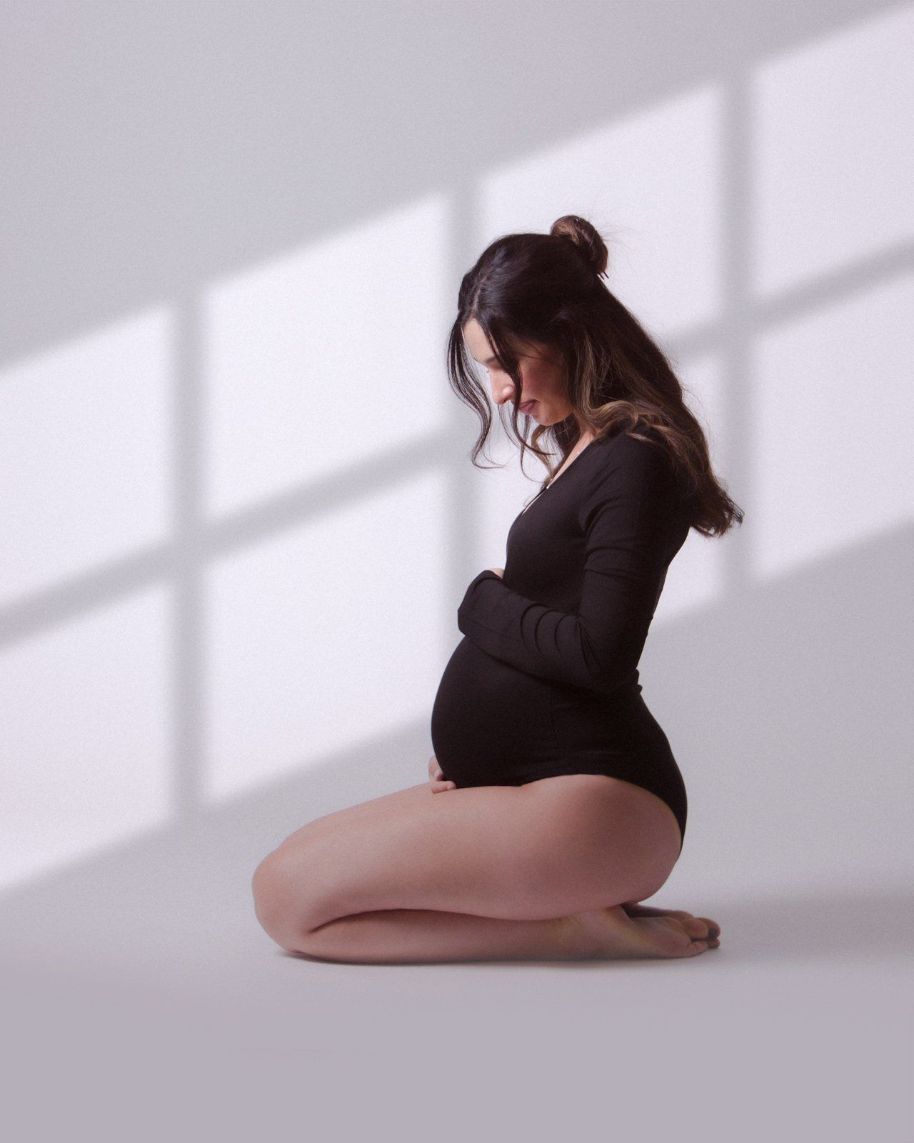 Pregnant woman in black bodysuit kneeling gracefully, cradling her baby bump in a softly lit studio.