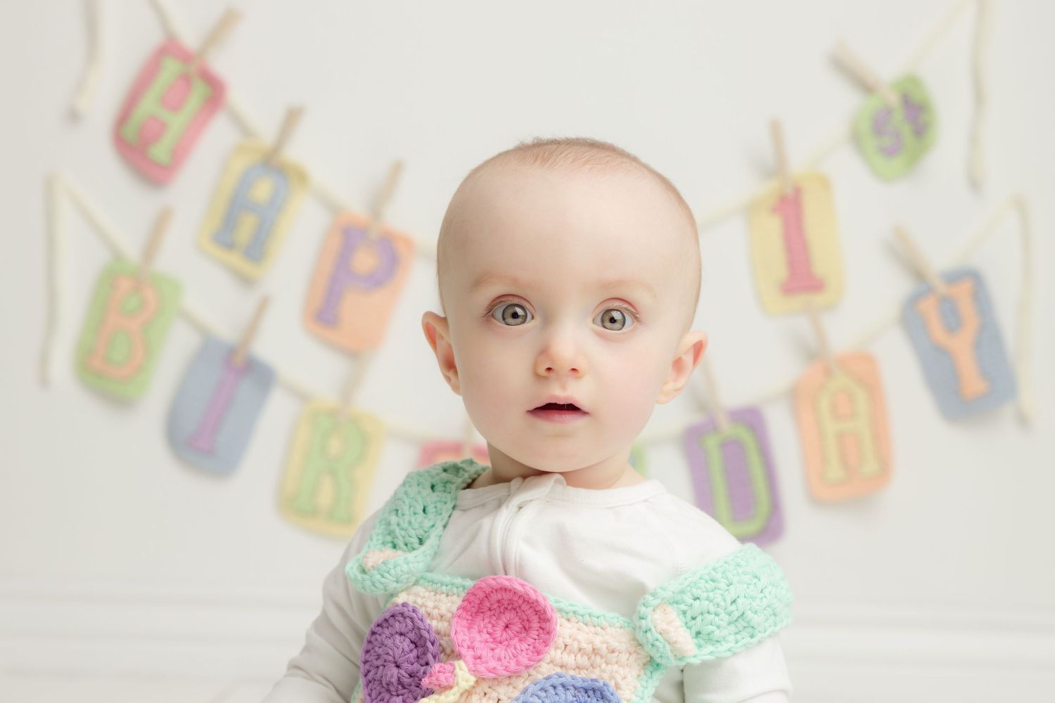 Baby wearing colorful knit sweater sits in front of wall decorated with pastel baby clothes and toys.