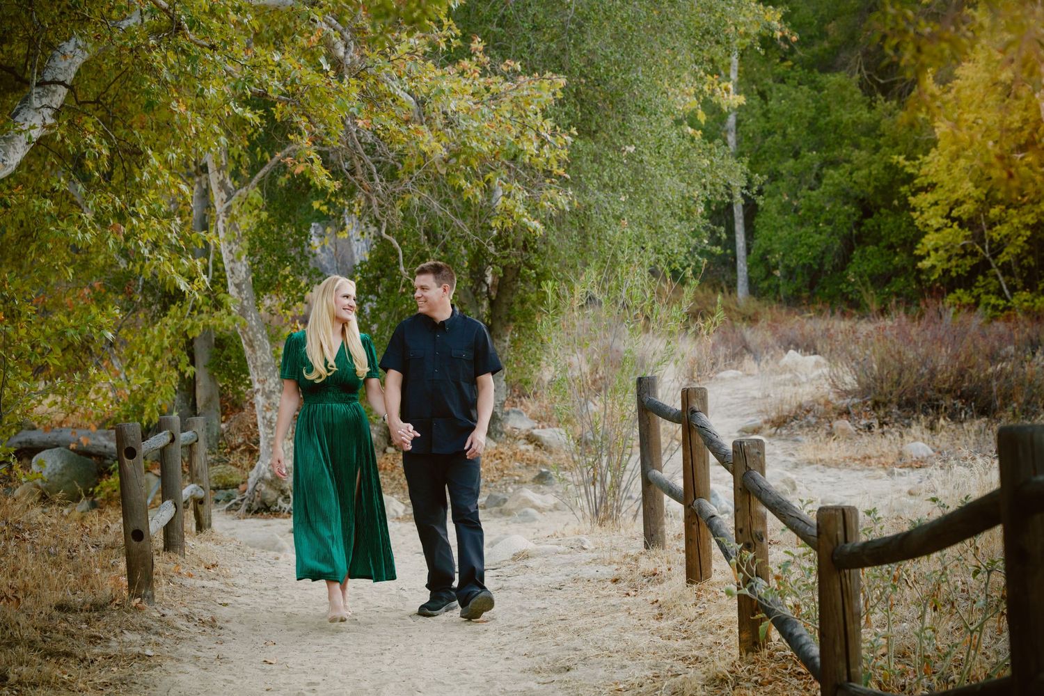 Couple walking hand in hand along a sunlit dirt trail lined with wooden fences, surrounded by autumn trees, the woman in a green dress.