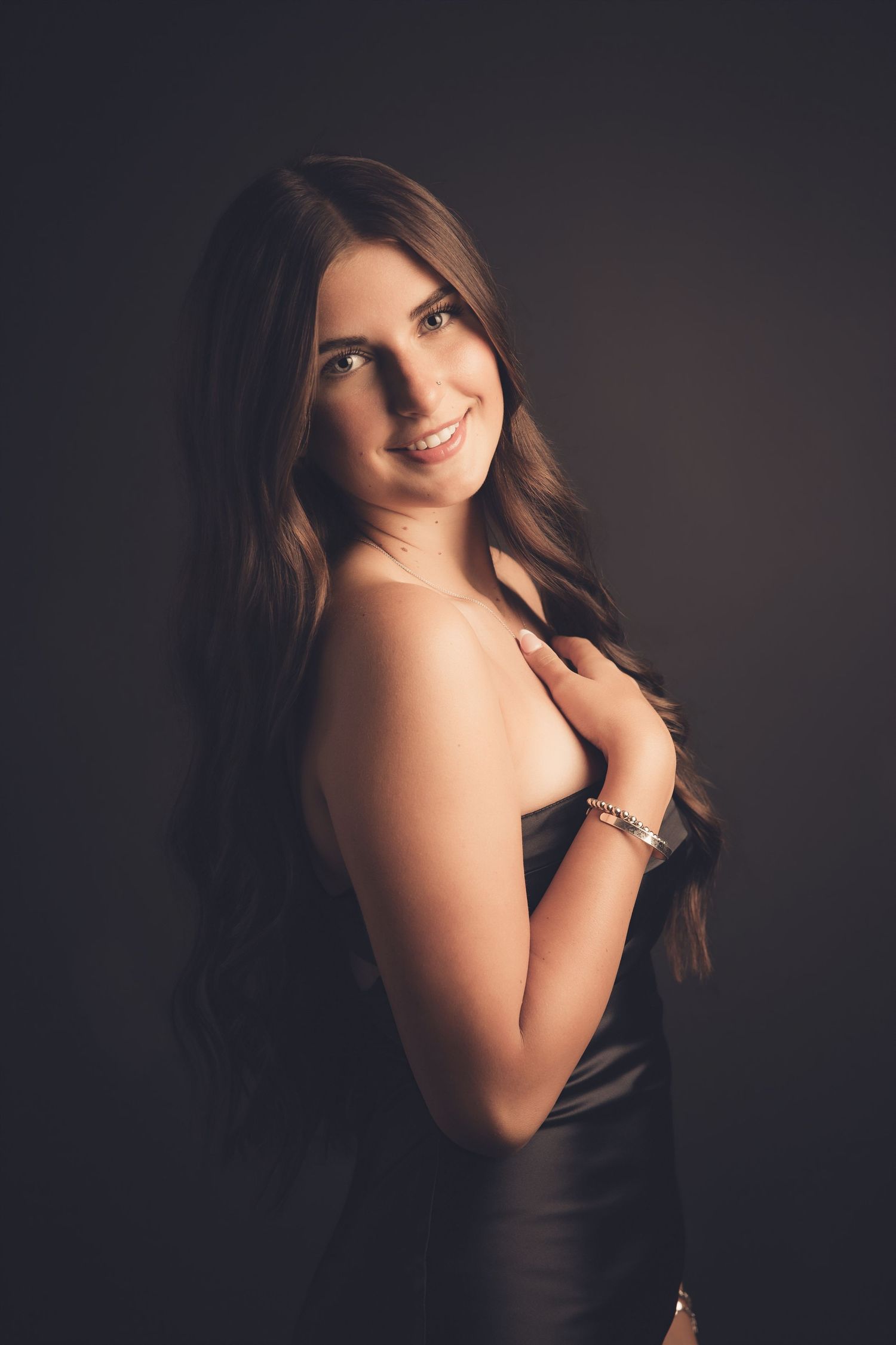 Elegant young woman with long brown wavy hair in a black strapless dress smiling over shoulder on dark background.