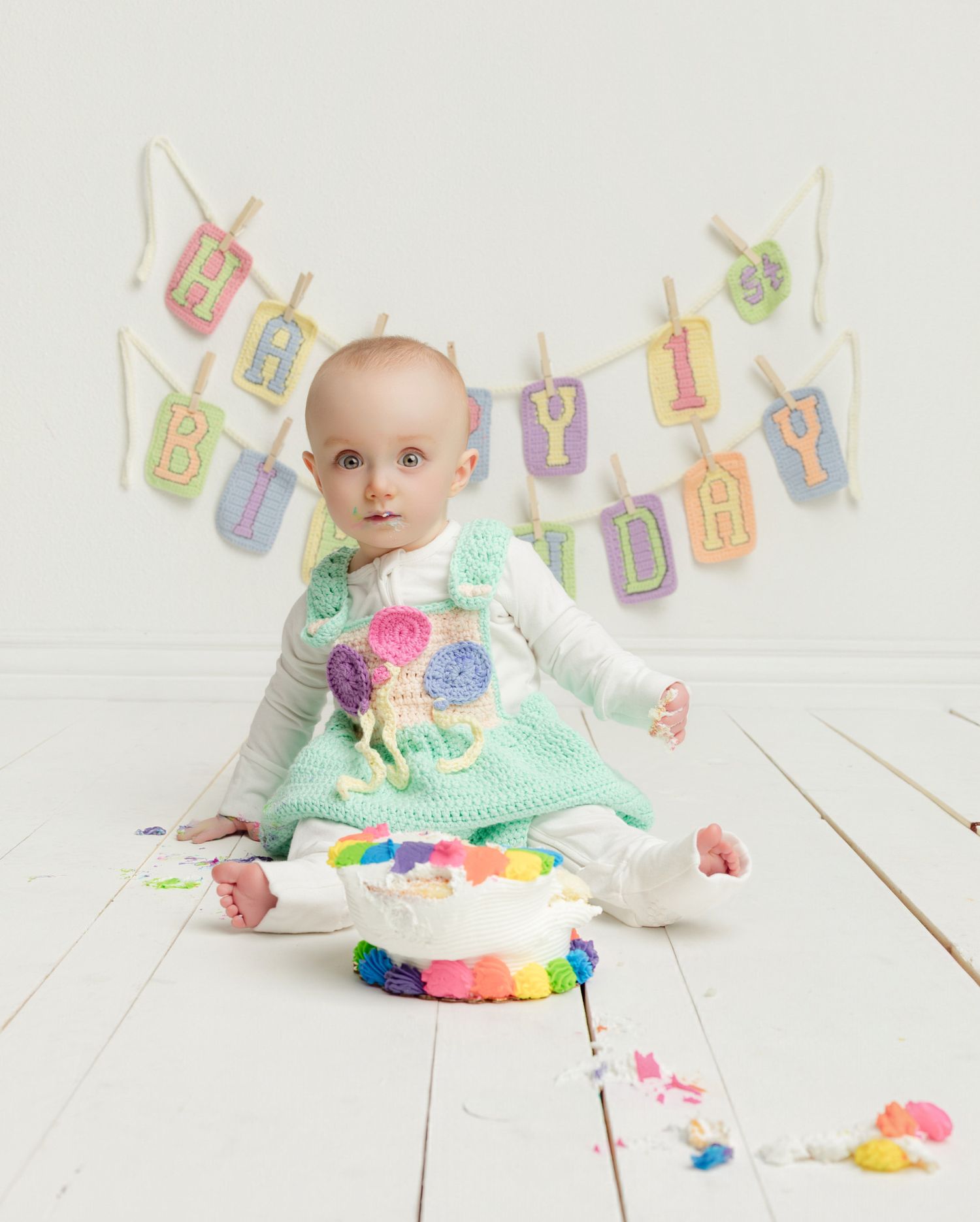 Baby in mint crochet dress sits beside rainbow smash cake under Happy 1st Birthday banner on white wood floor.