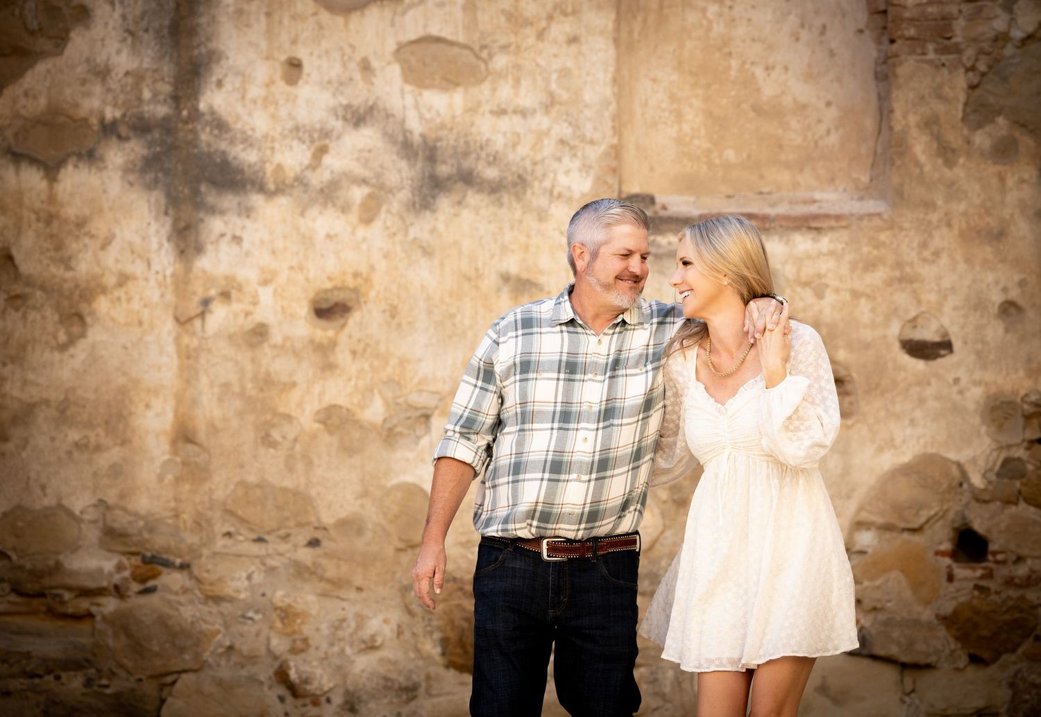 A couple shares a romantic moment against a rustic stone wall while dressed casually for an outdoor photoshoot.