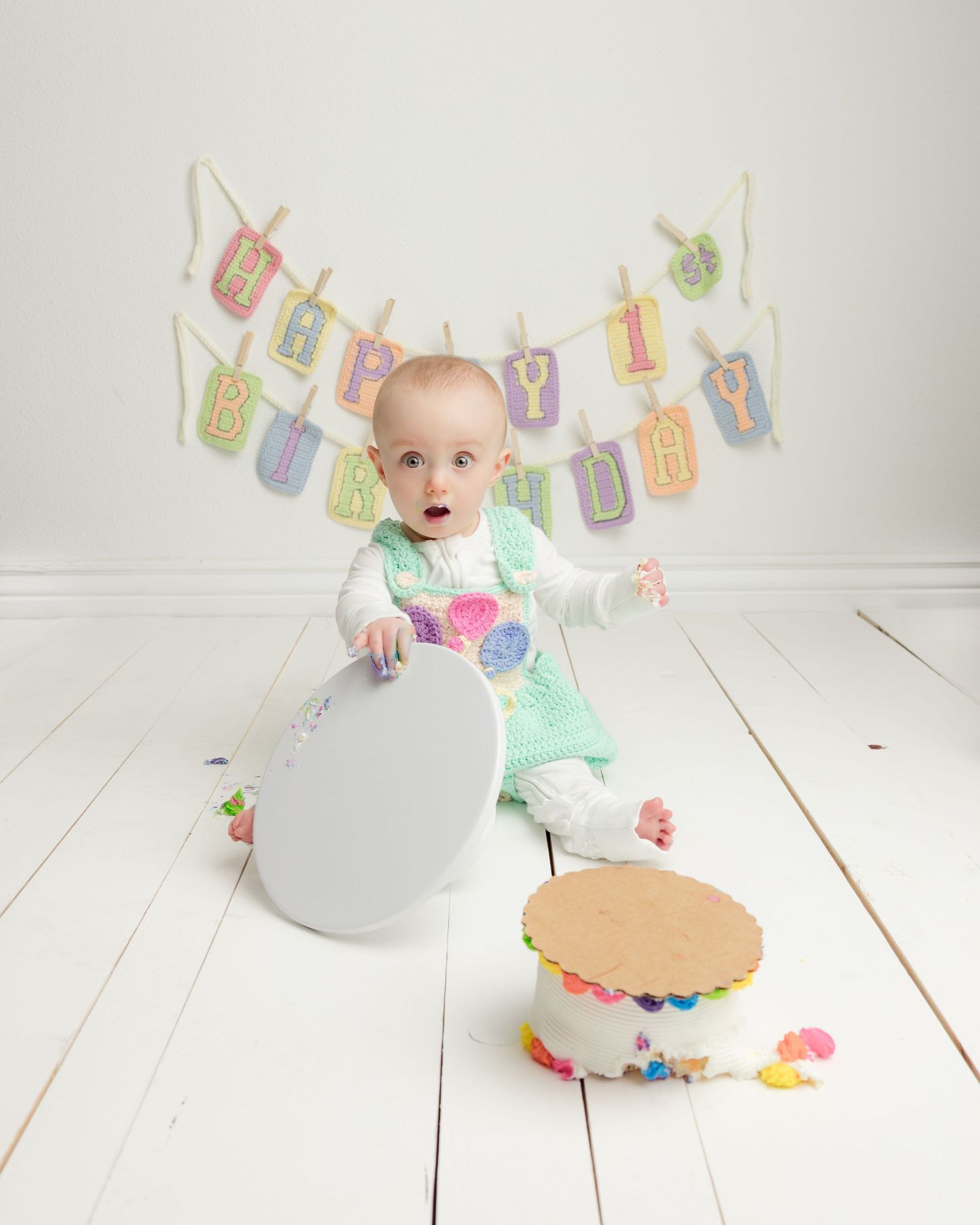 Baby in mint green outfit sits on white wooden floor with birthday decorations and cake smash setup.