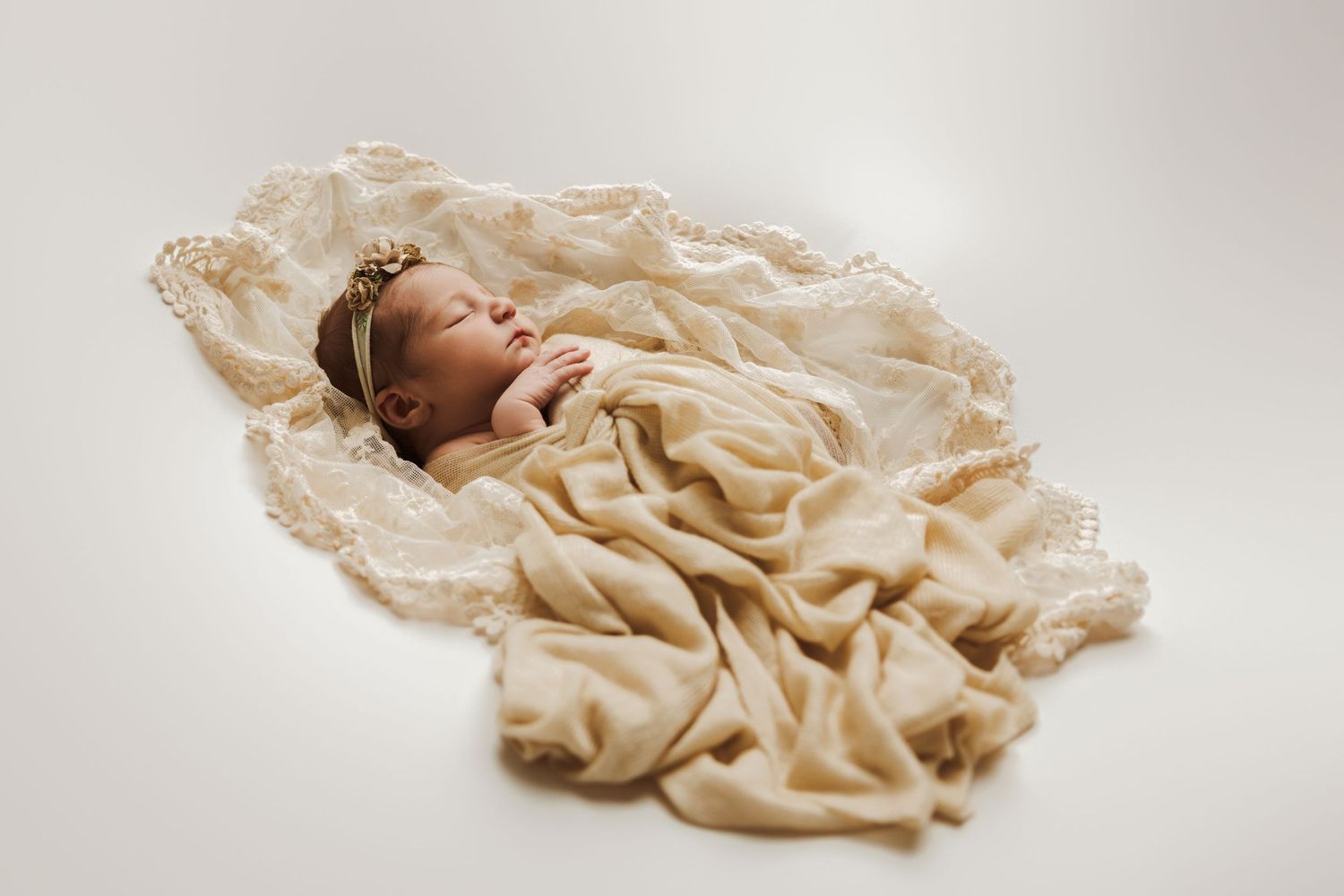 A sleeping baby wrapped in cream-colored fabric lies peacefully in a soft, minimalist studio setting.