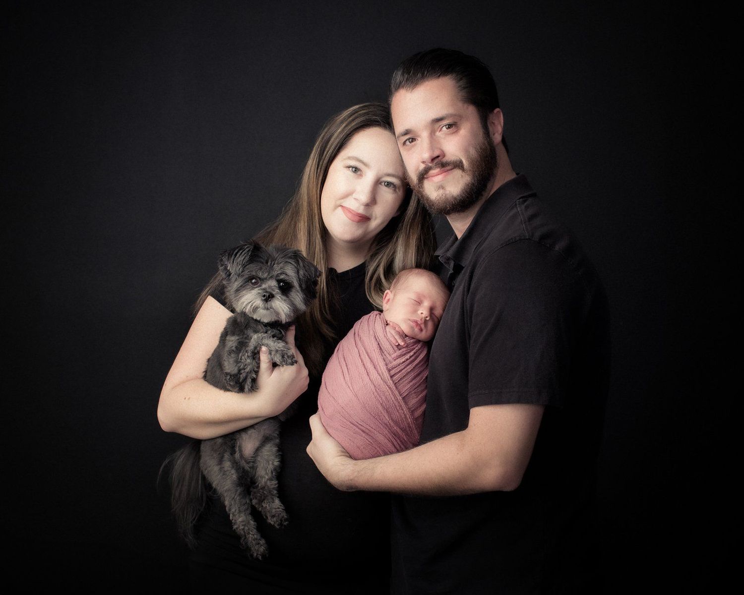 A family poses for a professional studio portrait against a dark background with their small gray dog and sleeping newborn.