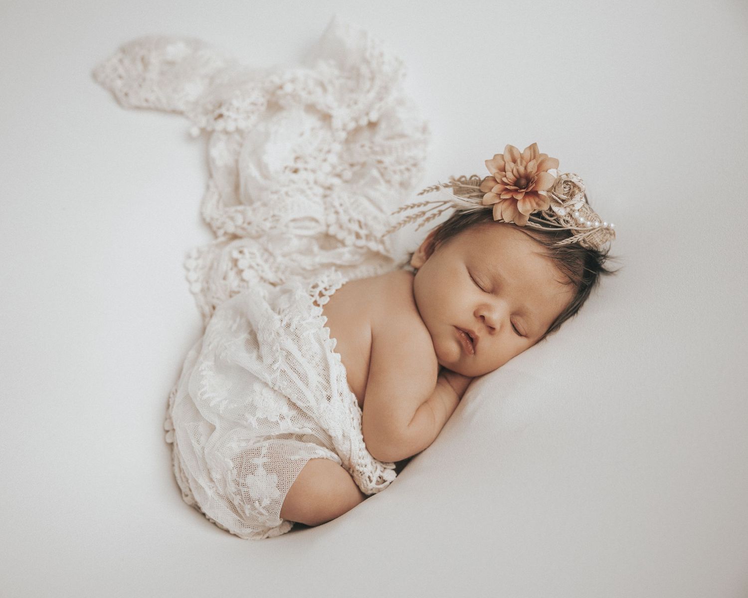 Serene newborn baby sleeping peacefully wrapped in delicate white lace against soft gray backdrop.