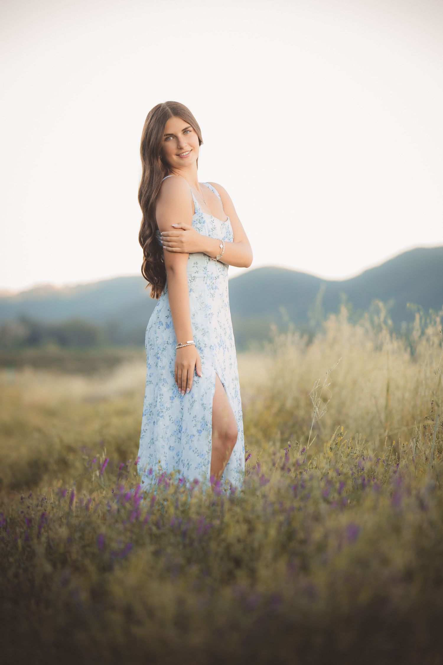 Young woman in floral blue maxi dress standing in wildflower field at golden hour with mountain backdrop.