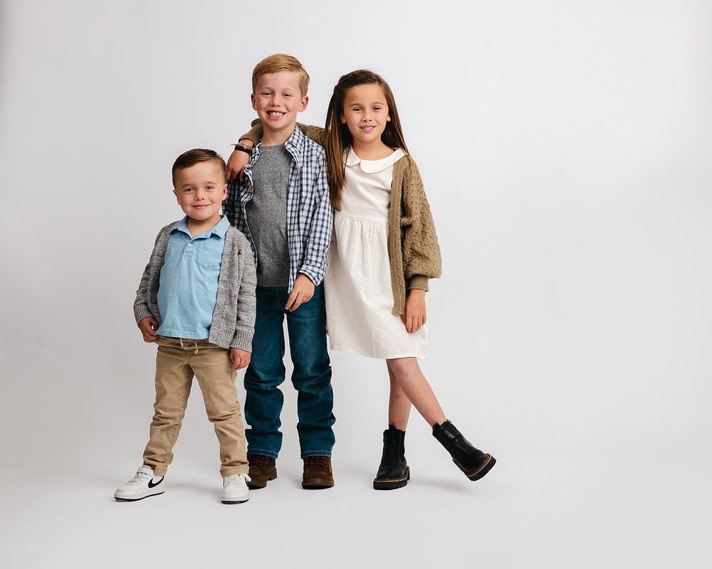 Three children stand together against a light gray background wearing casual fall clothing and boots.