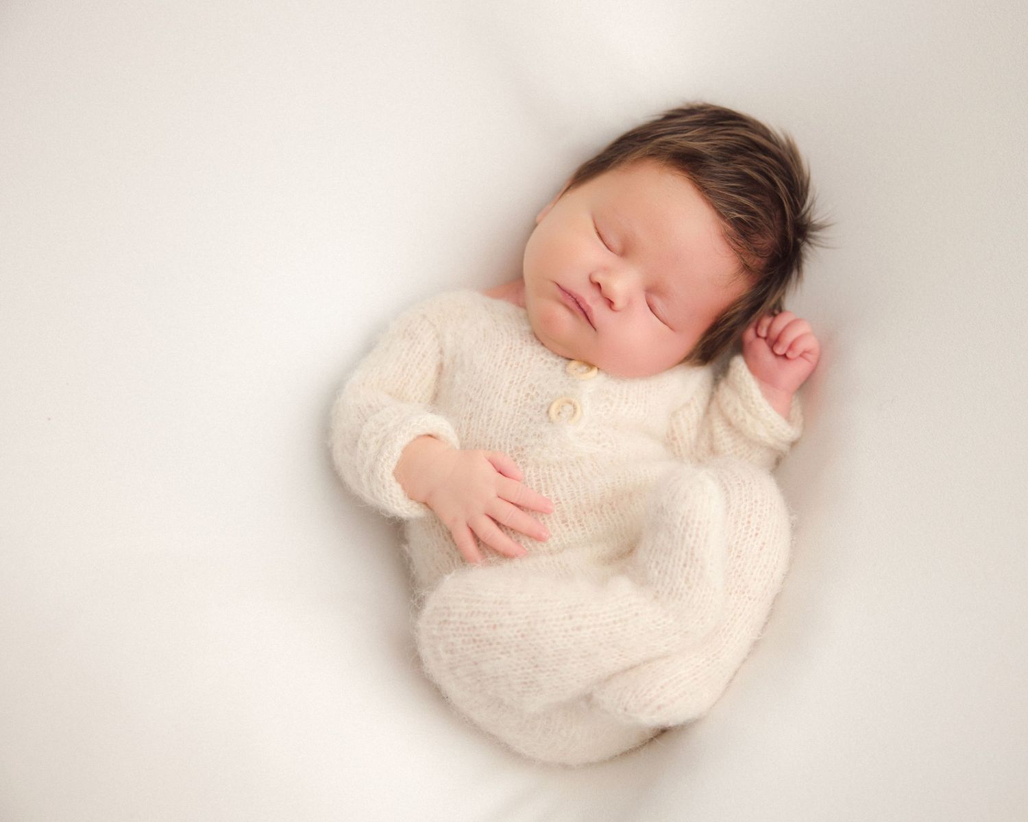 A sleeping baby wearing a soft cream-colored sweater lies peacefully on a white backdrop.