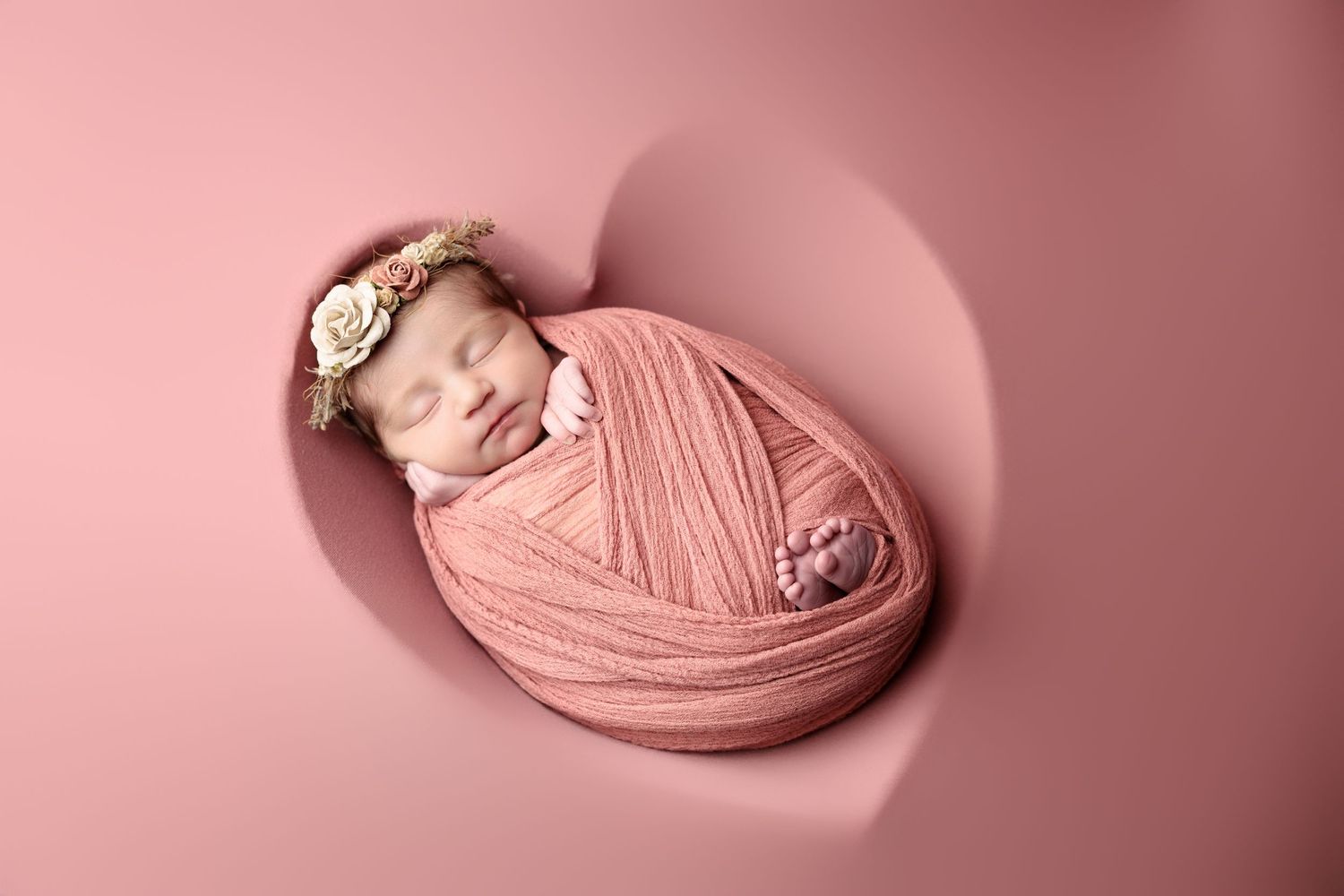 Newborn baby peacefully sleeping while wrapped in soft pink fabric on heart-shaped cushion.