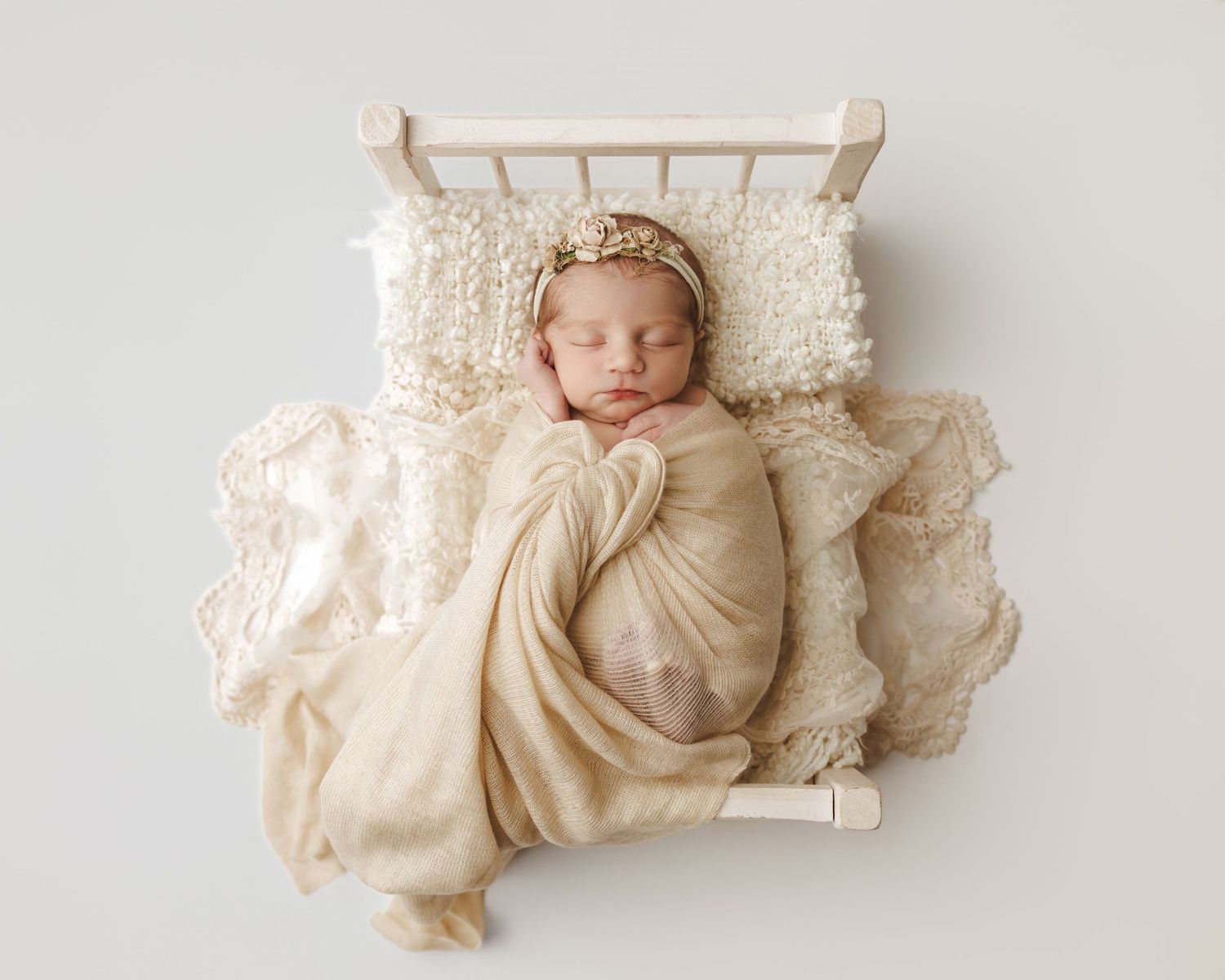 A sleeping baby wrapped in cream-colored fabric lies on white lace pillows with a delicate crown headpiece.