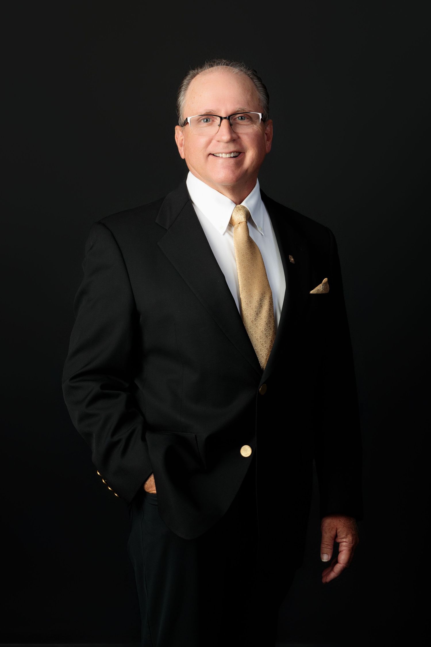 Professional portrait of a person in a black suit and gold tie against a dark background.
