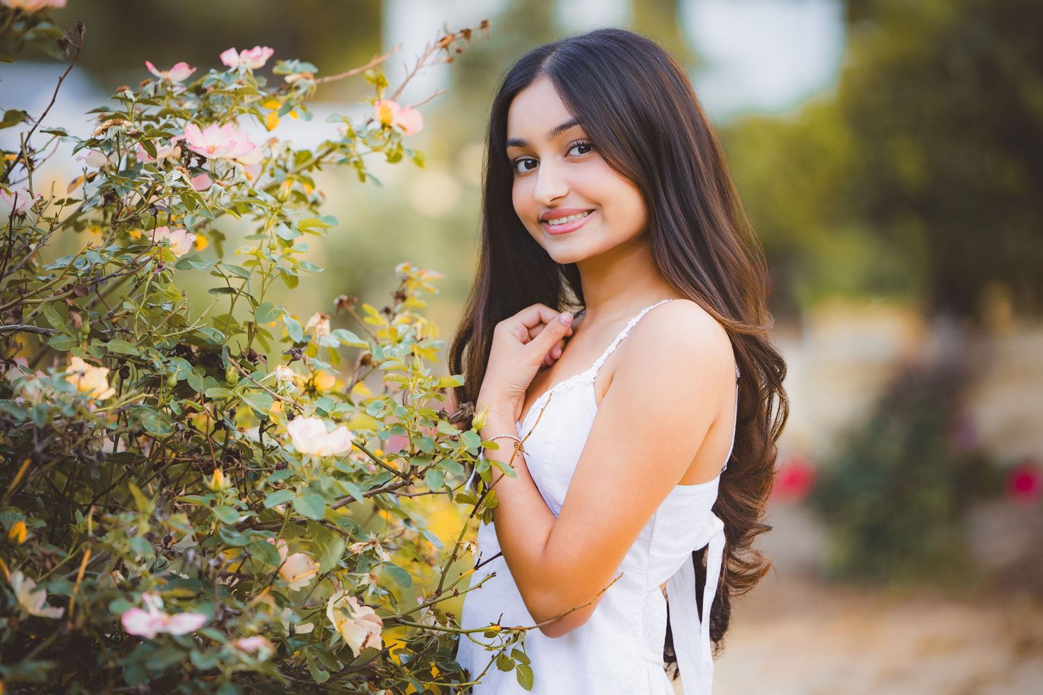 Young woman in white dress smiling beside blooming rose bushes in a sunlit garden.