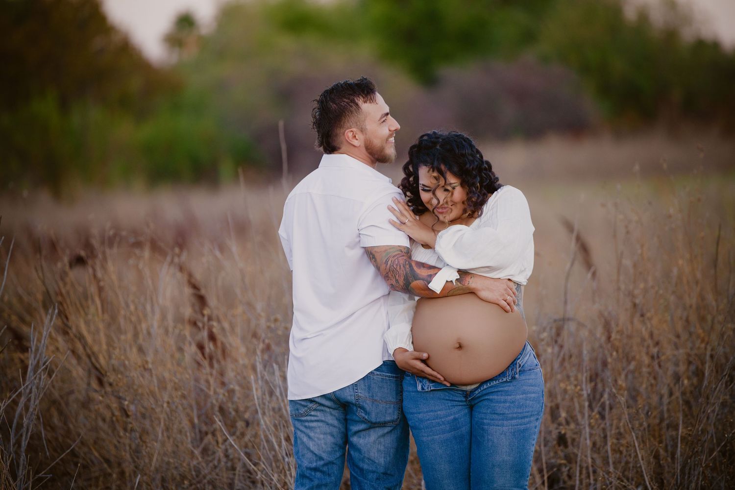 Couple embracing in a field during a maternity photoshoot, woman showing baby bump, wearing white tops and jeans.