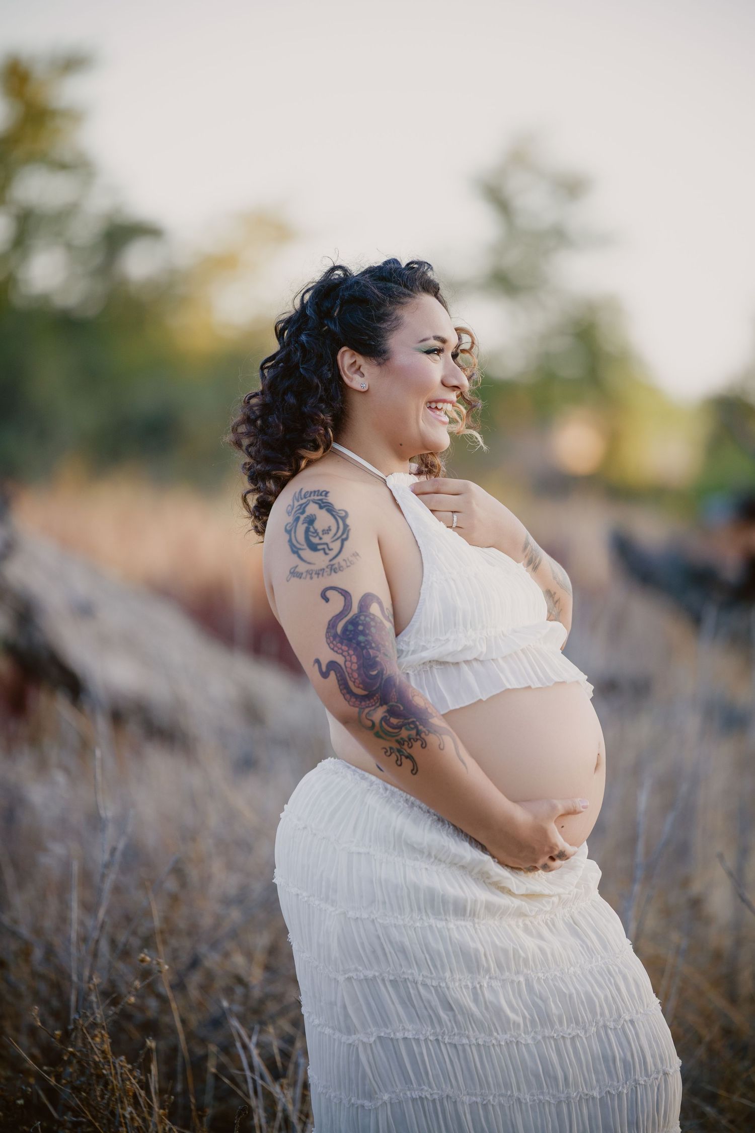 Smiling pregnant woman with tattoos in white dress cradling baby bump outdoors in golden field at sunset.