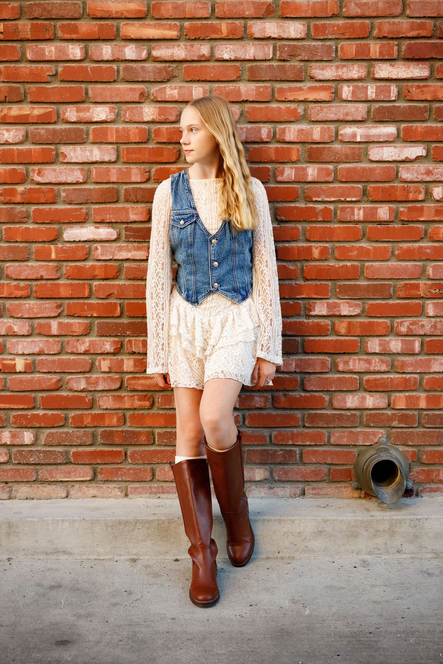 A person in a denim vest, lace dress and brown riding boots poses against a red brick wall.