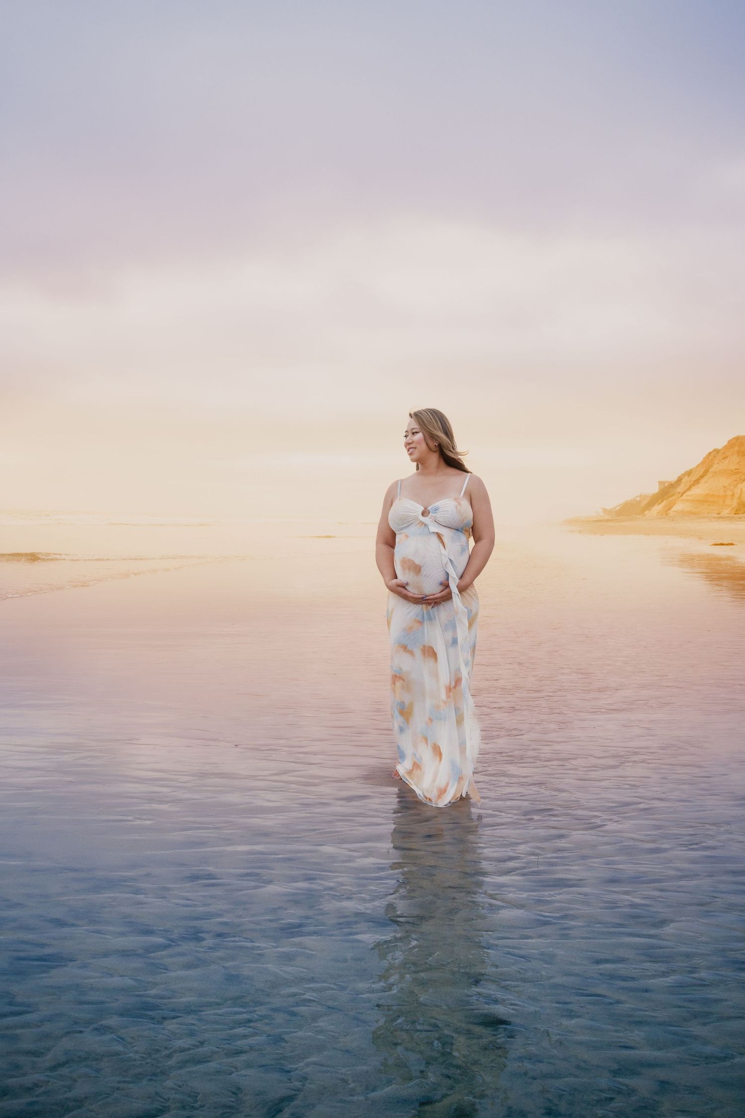 Pregnant woman in floral dress standing in shallow ocean water at sunset, cradling her baby bump on a serene beach.