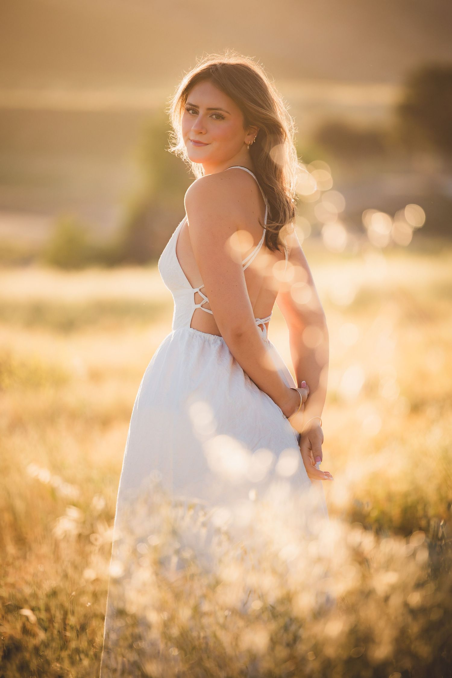 Young woman in white sundress standing in golden sunlit field at sunset.