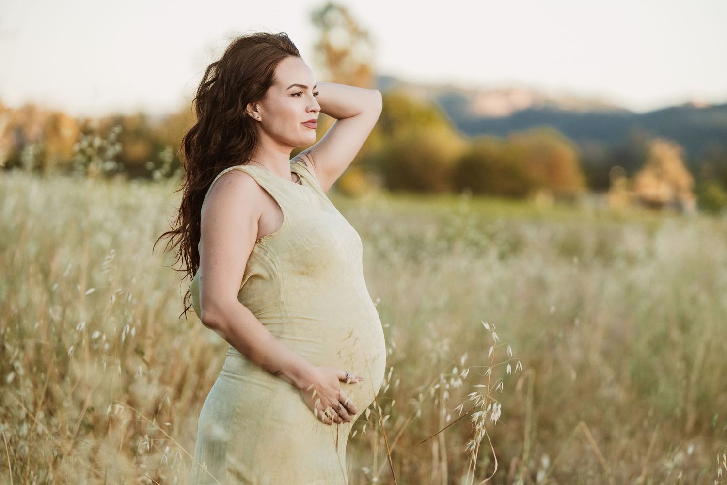 Pregnant woman in yellow dress standing in golden field at sunset, cradling baby bump with mountains in background.