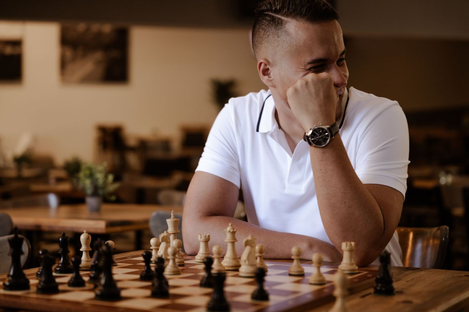 Man in white polo shirt deeply focused on a chess game, resting chin on hand, wearing a watch.