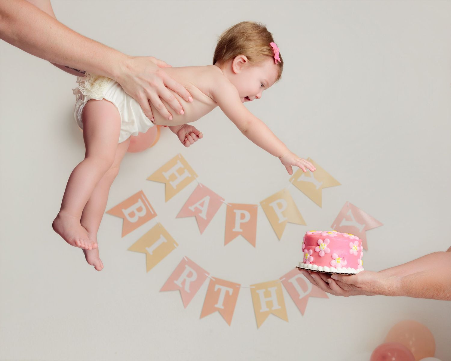 Baby reaching for a pink birthday cake under a Happy Birthday banner, held by adult hands in a fun first birthday photo.