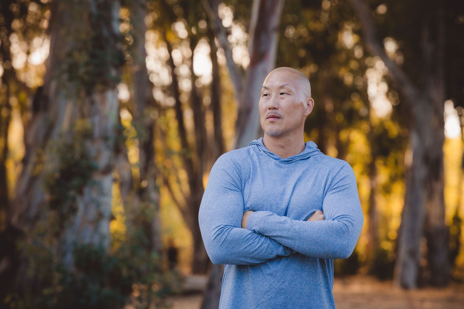 Person in light blue sweater standing with arms crossed in a wooded area during golden hour sunset.