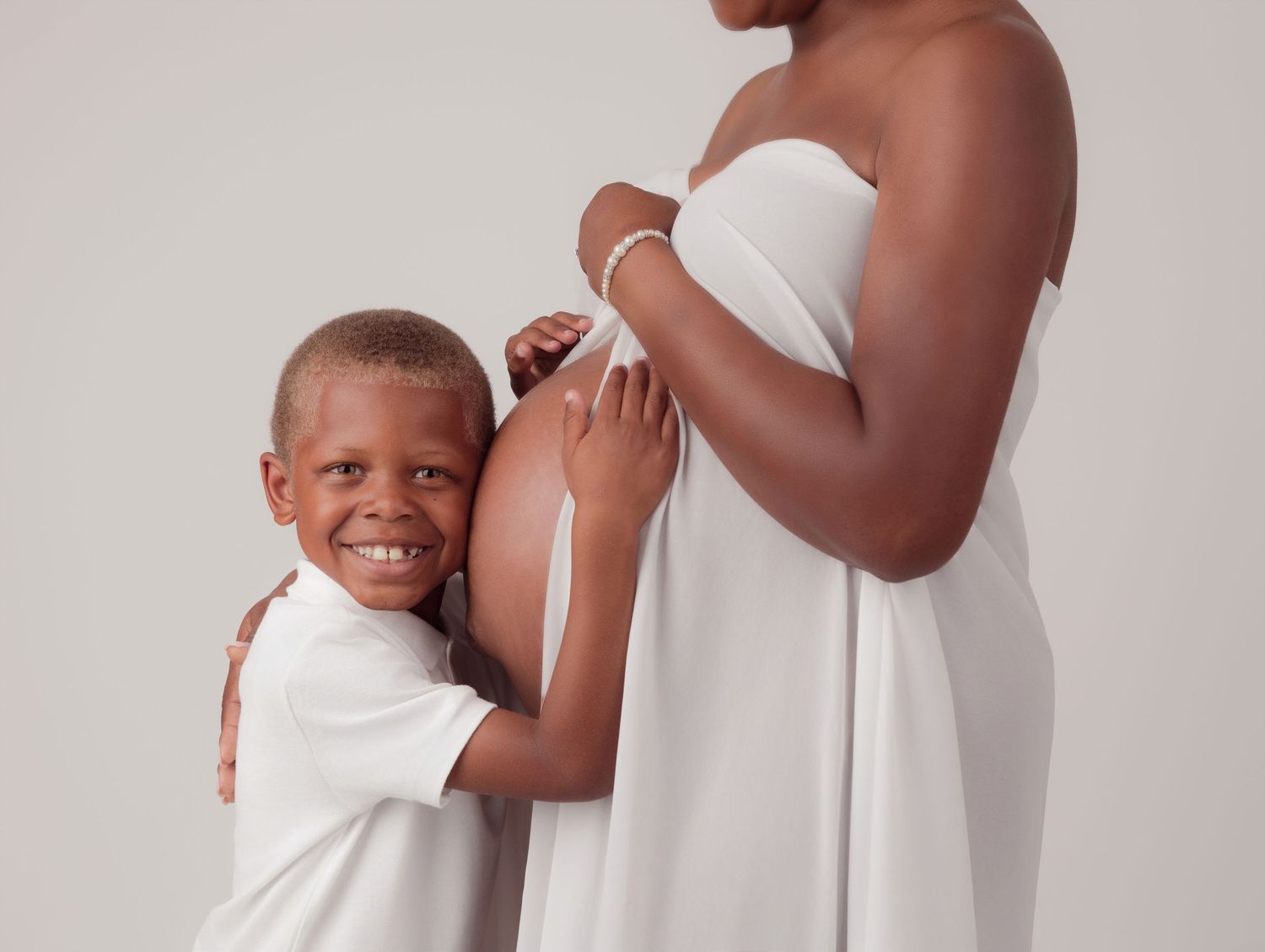 A person in a white dress stands while someone in white embraces them in a warm moment against a light background.