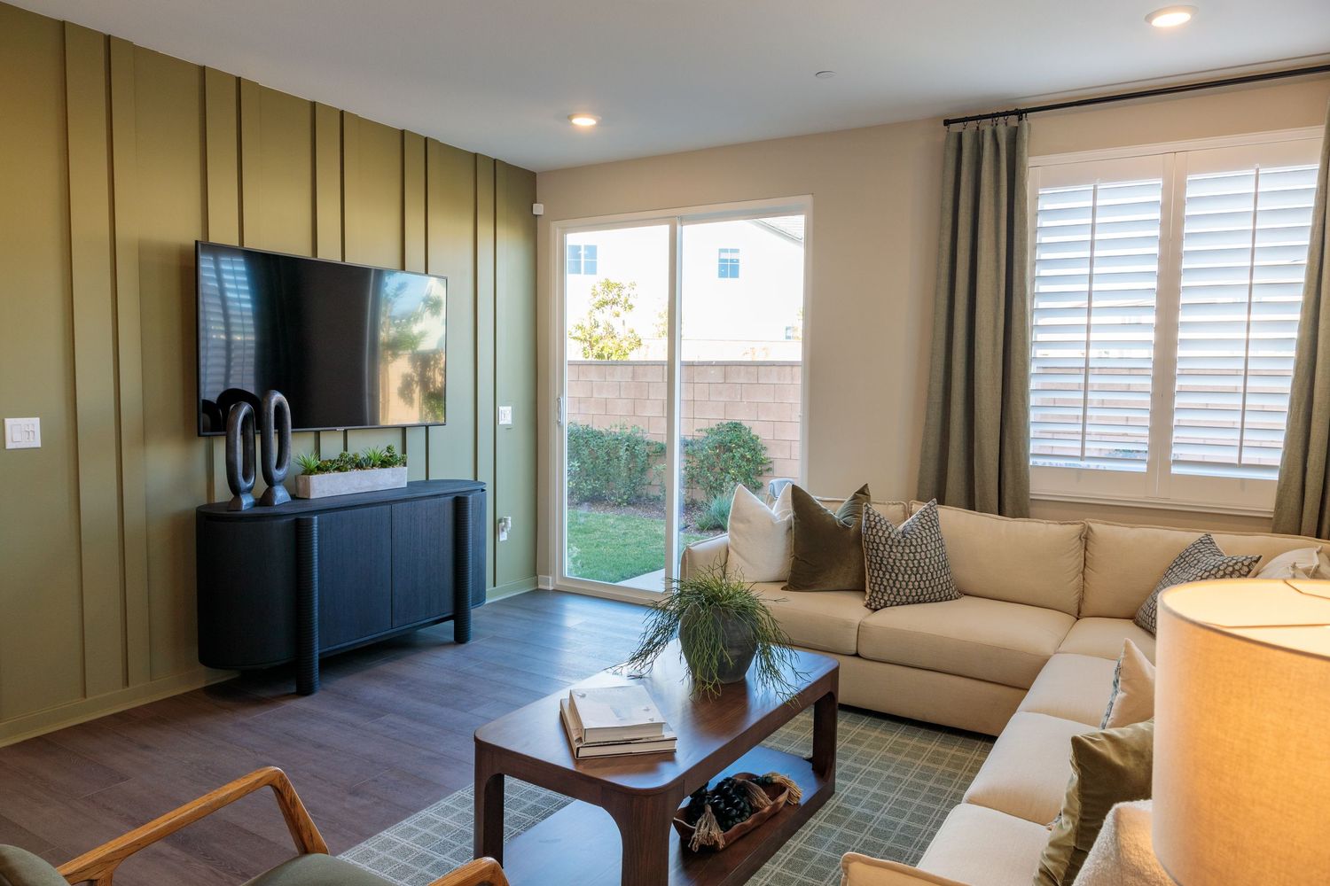 A modern living room features sage green walls, beige sectional sofa, and large windows with white plantation shutters.