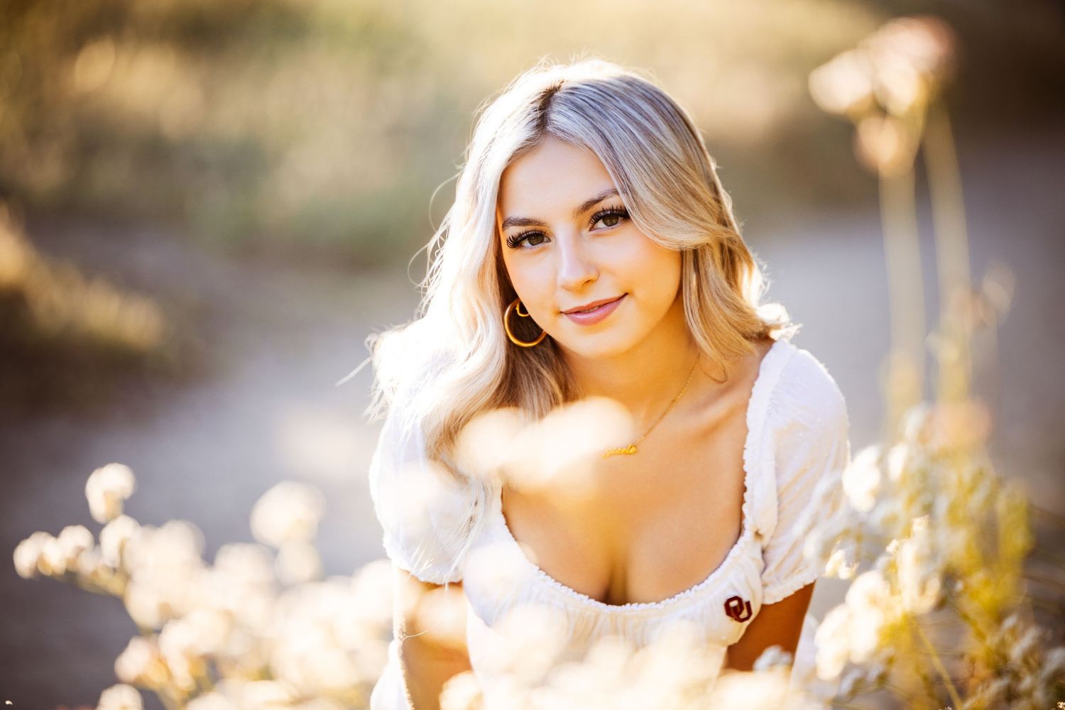 Blonde woman in white top posing outdoors in golden sunlight among wildflowers for a portrait photo.