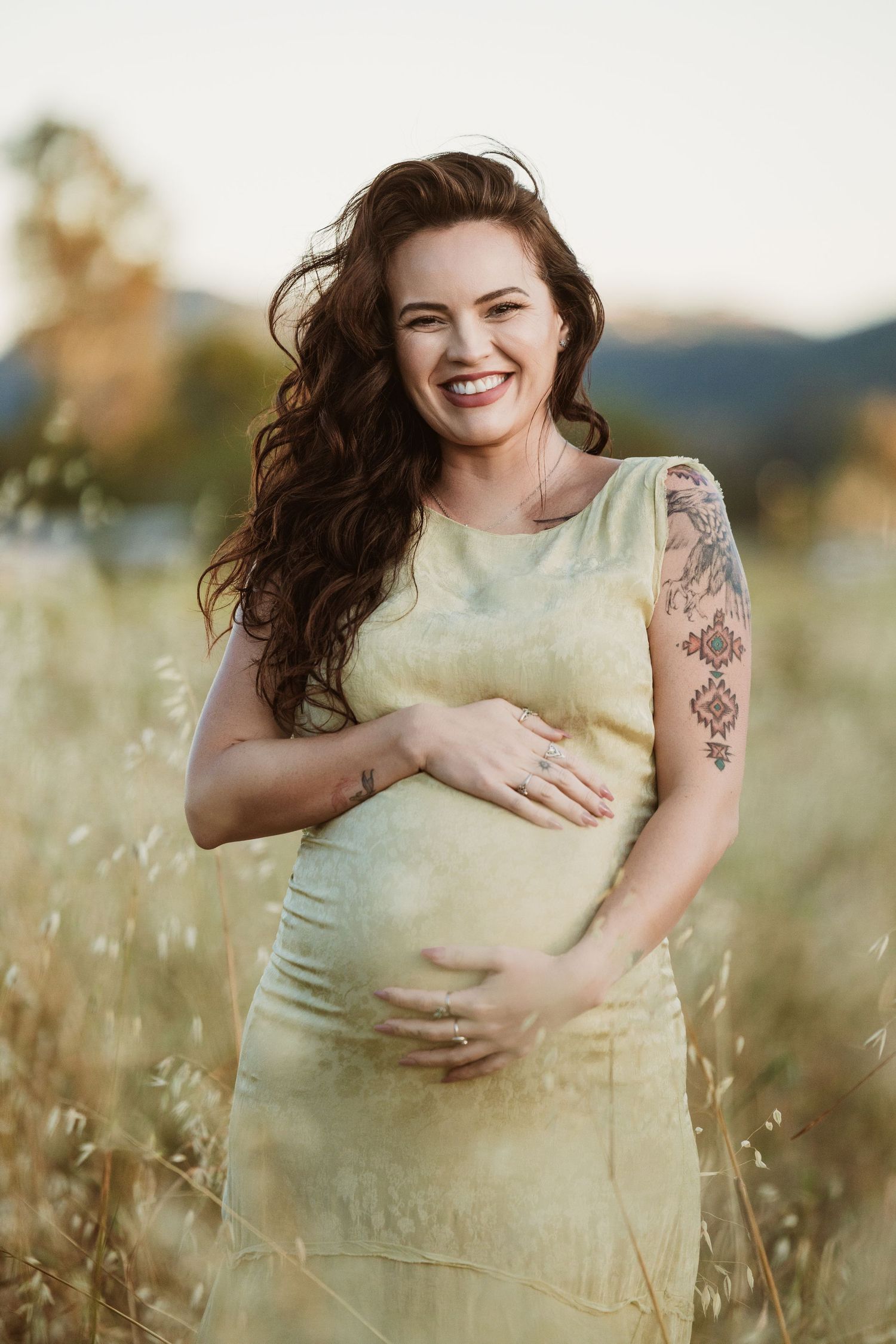 Smiling pregnant woman in yellow dress holding baby bump in golden field, with tattoo on arm and long curly brown hair.