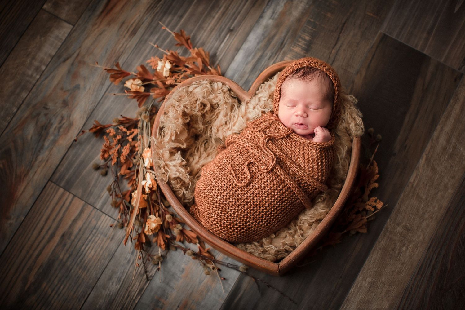 Sleeping newborn wrapped in brown knit swaddle lying on rustic wooden floor with autumn leaves.