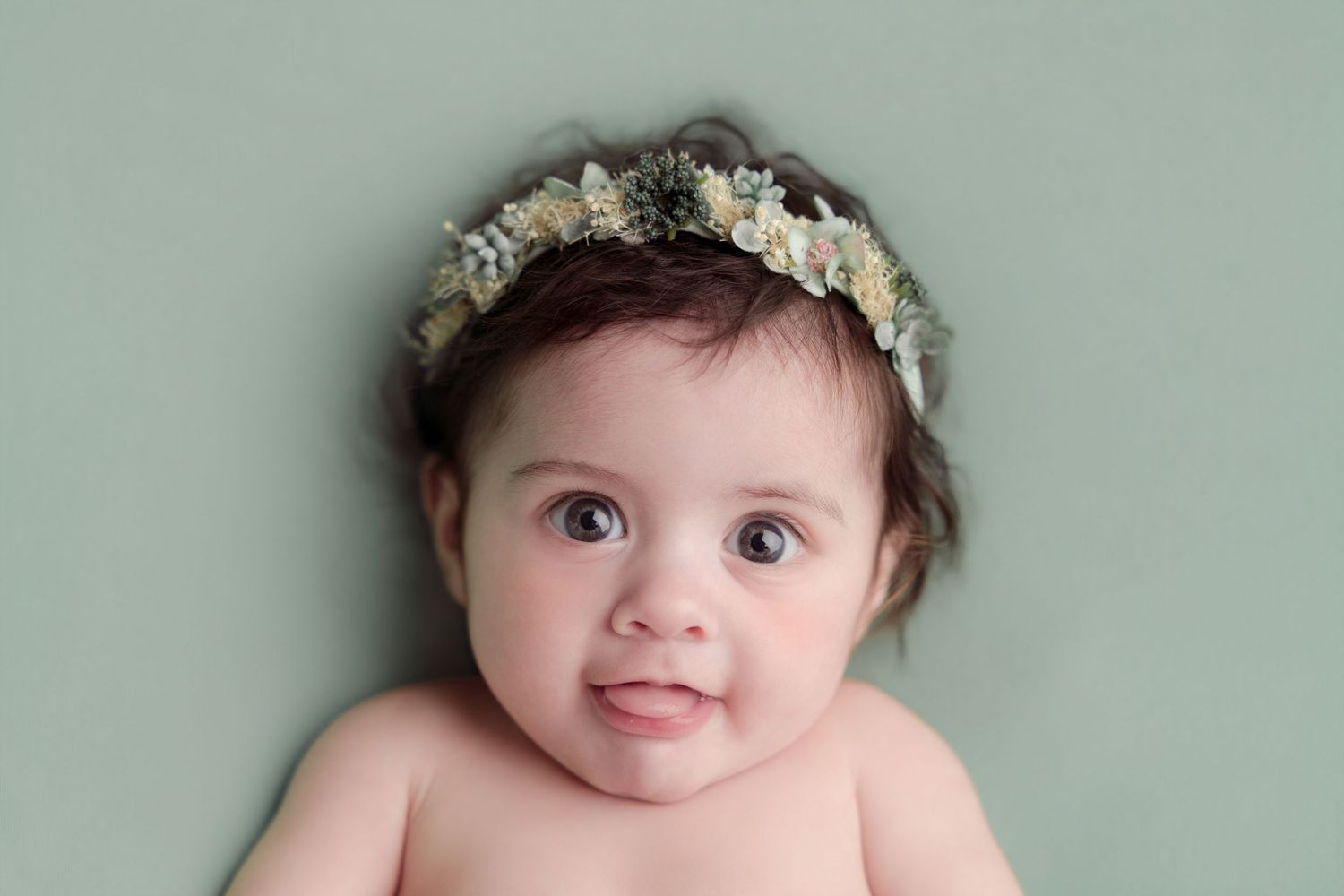 A baby wearing a delicate floral crown poses against a sage green background.
