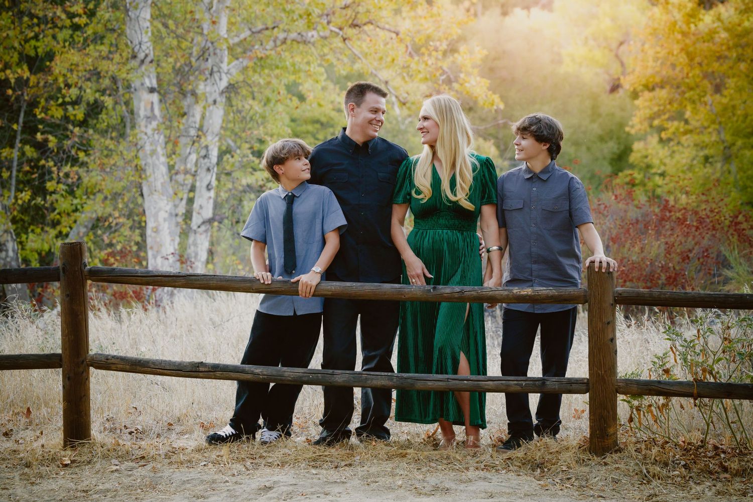 Family of four posing by a rustic wooden fence in an autumn park, with a woman in a green dress and three smiling males.