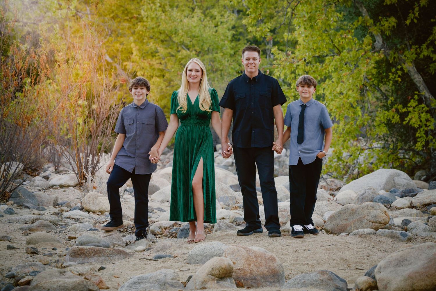 Family of four holding hands and standing on a rocky riverbed, with a woman in a green dress, a man and two boys in autumn light.