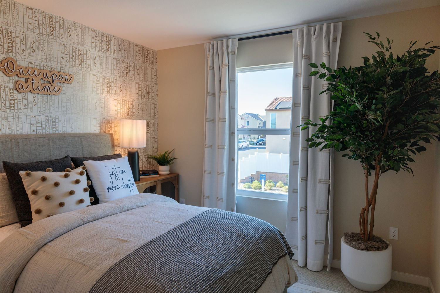 Modern bedroom interior with gray bedding, decorative pillows, potted plants and natural light from windows.
