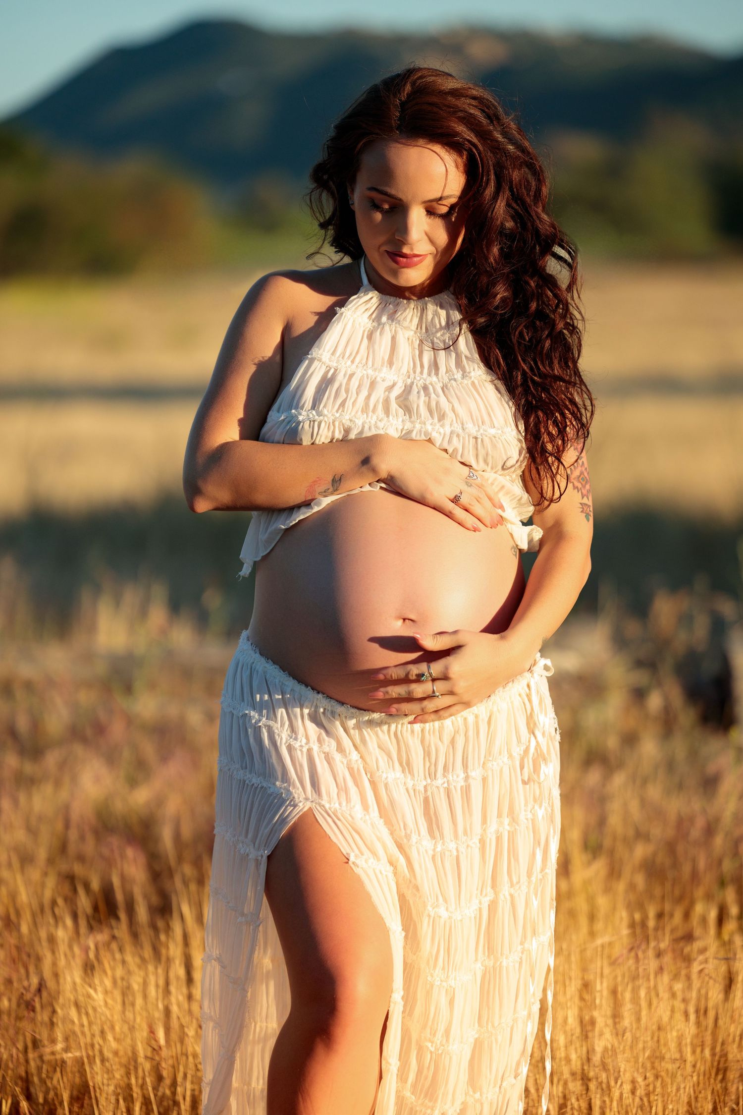 Pregnant woman in white dress cradling bare baby bump in golden field with mountain backdrop at sunset.