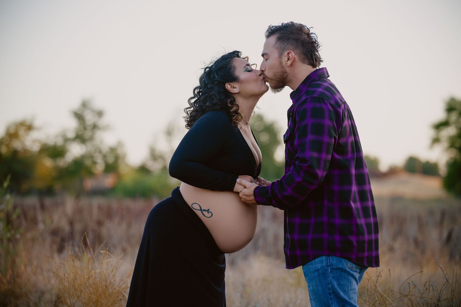 Pregnant couple sharing a tender kiss outdoors in a golden field during a maternity photo session at sunset.