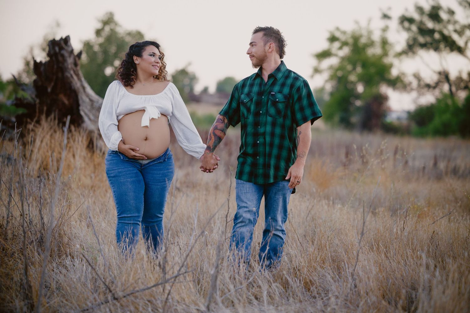 Pregnant couple holding hands walking through golden field, woman showing baby bump, man in plaid shirt, outdoor maternity photo.