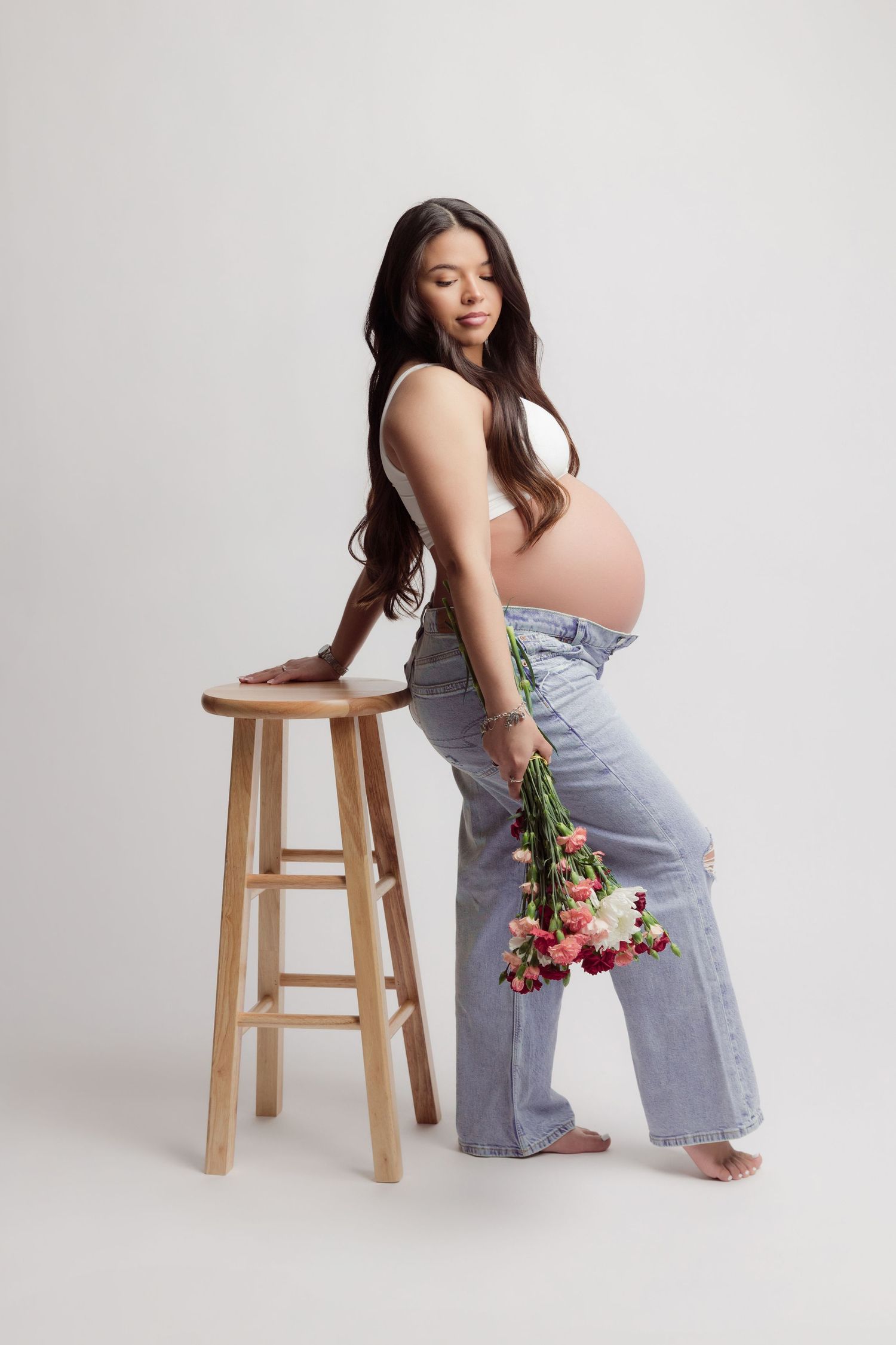A person in light blue jeans poses with pink tulips against a white backdrop in a minimalist studio setting.