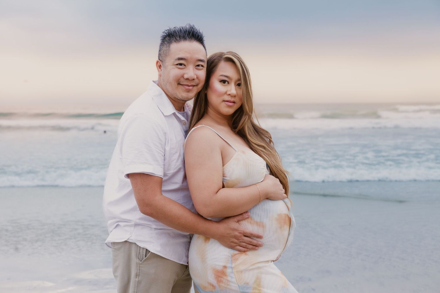 Couple posing on beach at sunset, woman in flowy maternity dress cradling baby bump, man embracing her from behind.