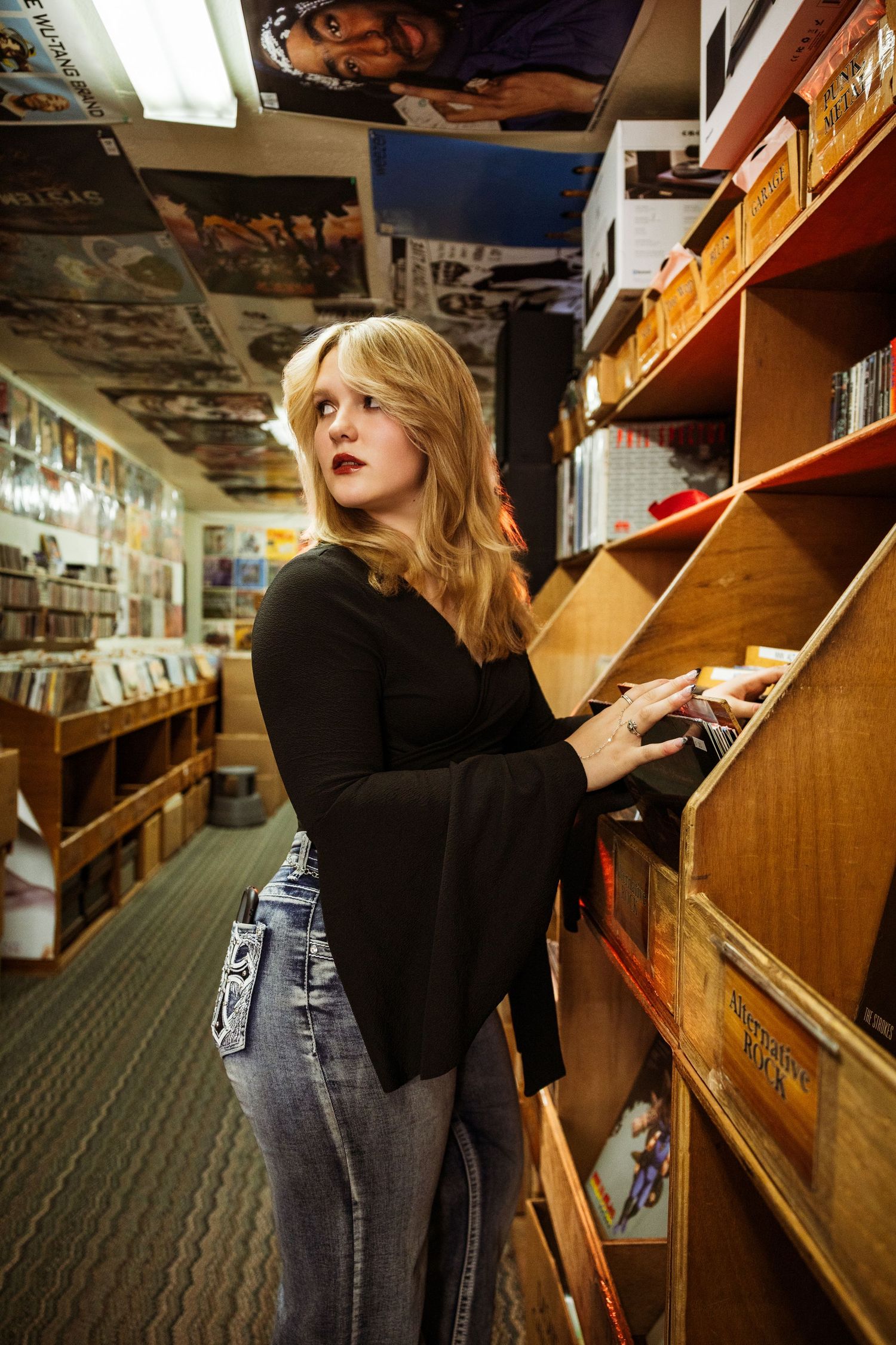 Young blonde woman browsing vinyl records in a cozy music store aisle, wearing a black top and gray jeans.