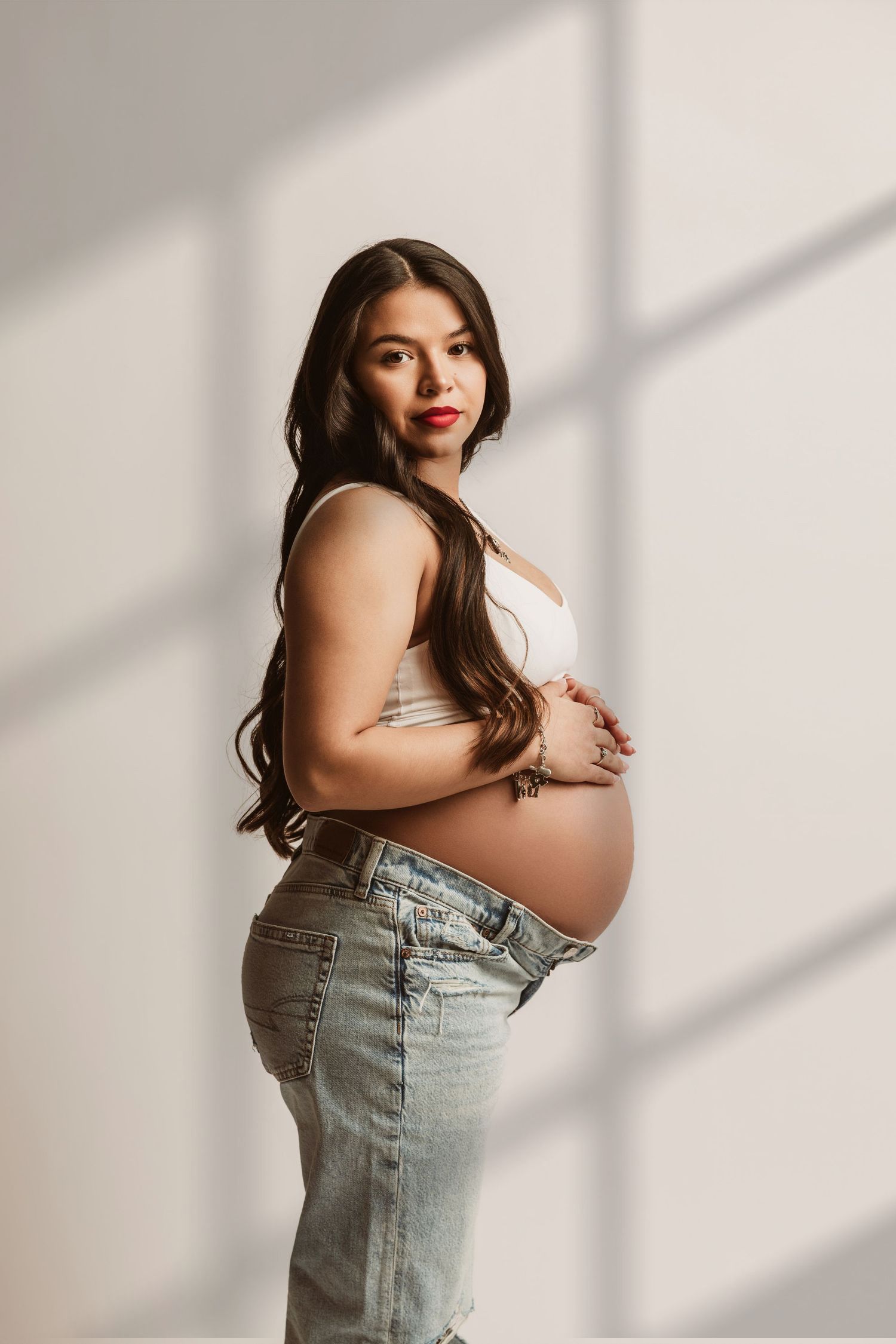 Pregnant woman in white crop top and jeans cradling baby bump, long dark hair, red lips, studio light.