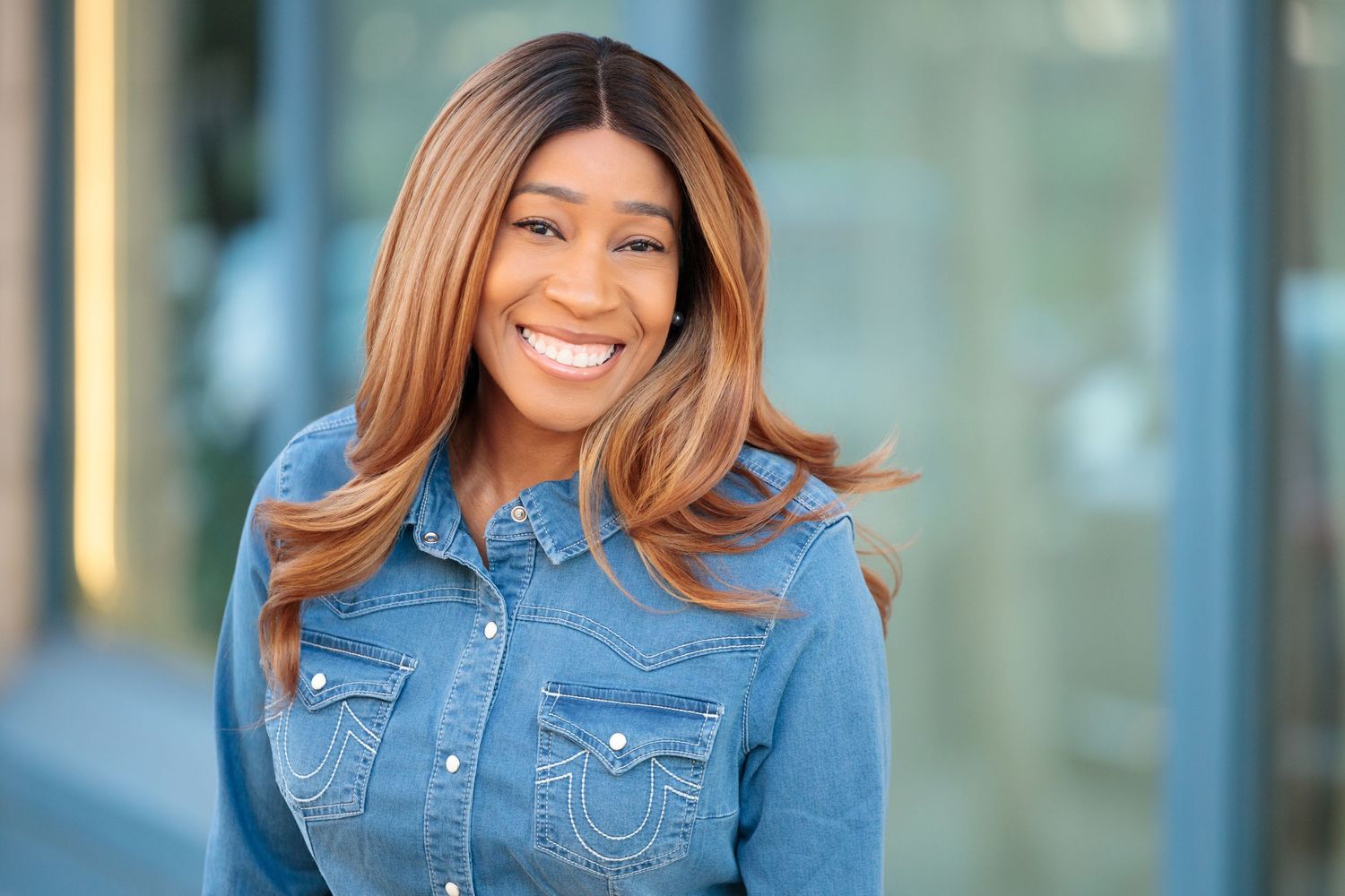 Smiling Black woman with long highlighted hair wearing a denim shirt, posing outdoors in natural light.