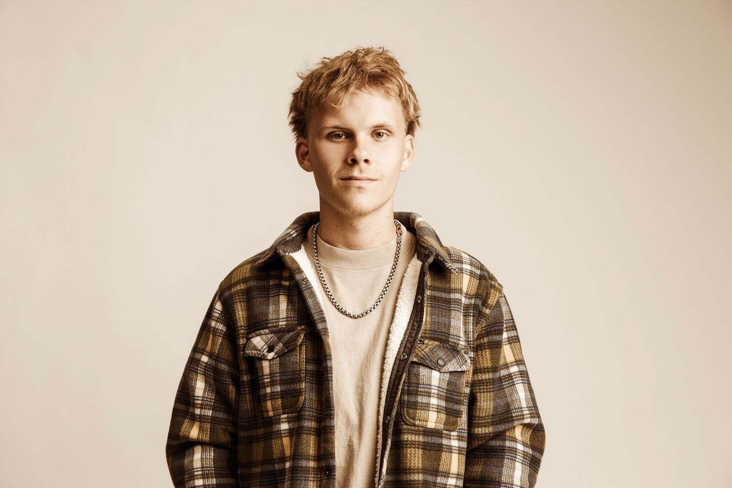 Young blond man wearing a plaid flannel shirt over a beige tee with a gold chain necklace posing against neutral background.