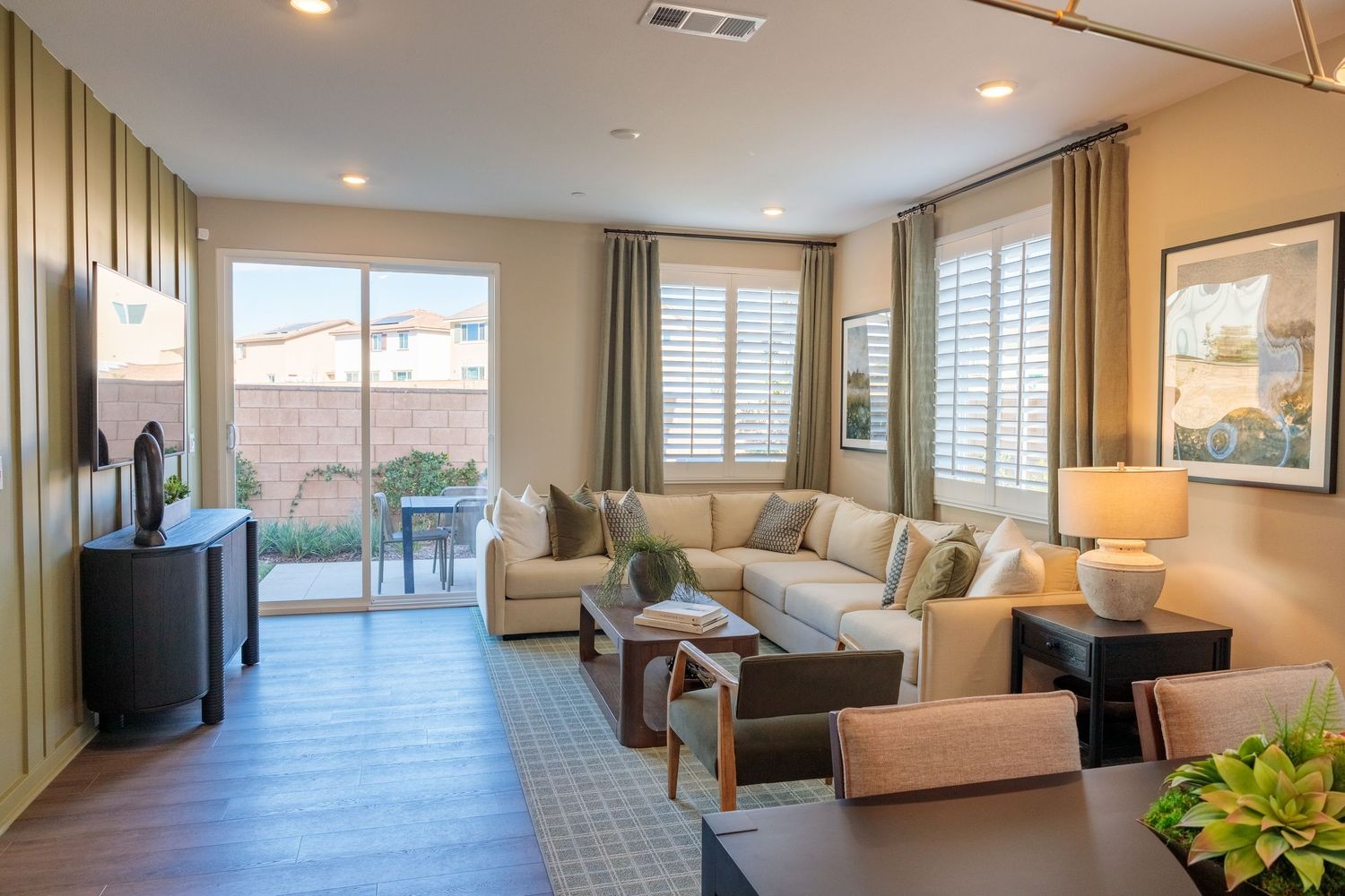 Modern living room with beige sectional sofa, hardwood floors, plantation shutters and sliding door leading to patio.