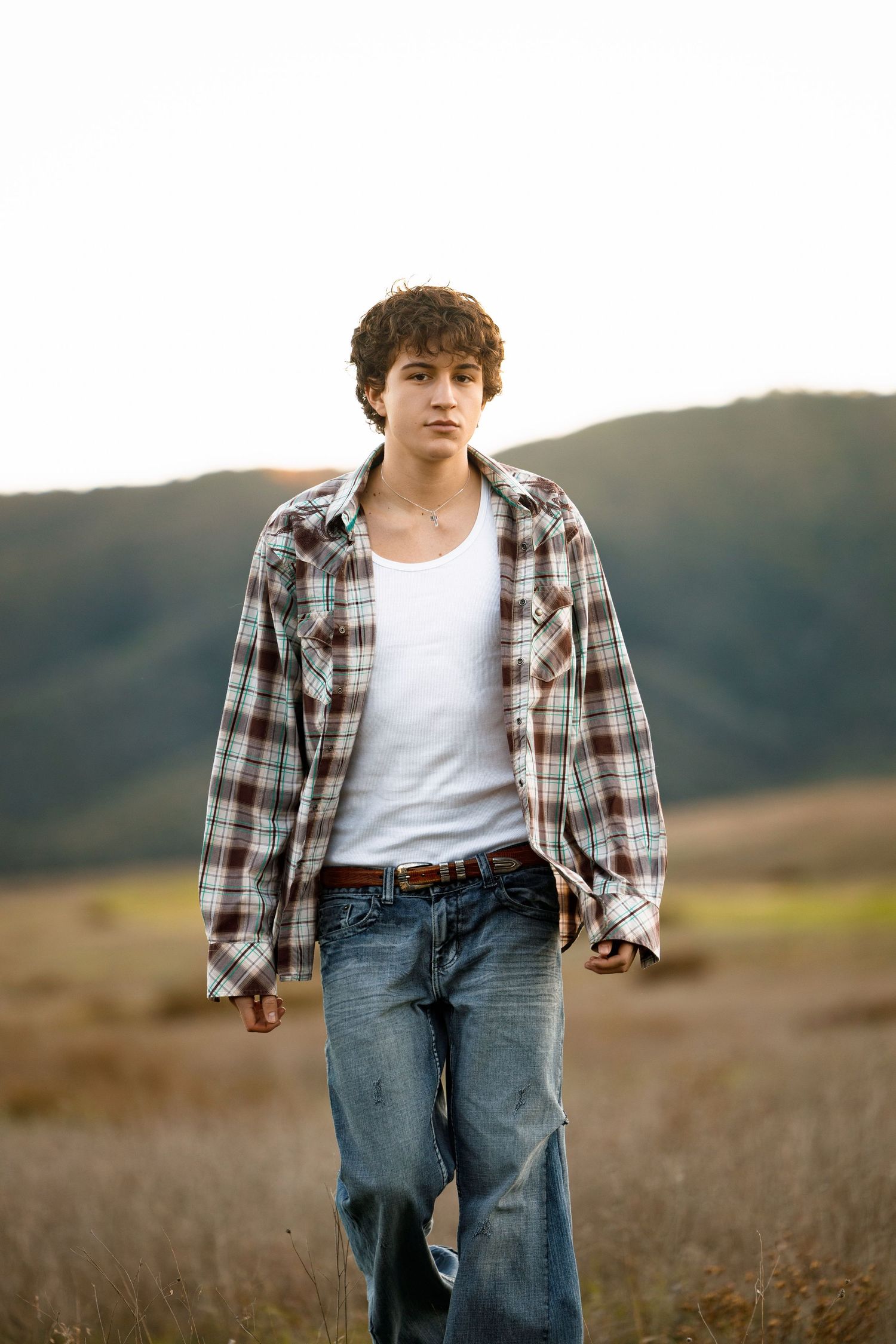 Young person in plaid shirt and jeans stands in golden field with rolling hills in background at sunset.